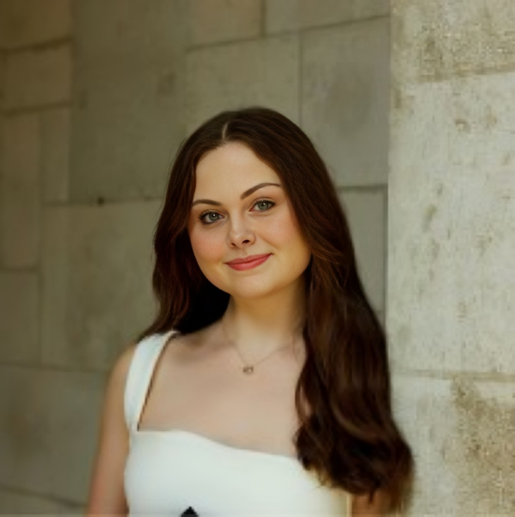 A young woman with long, wavy brown hair, light skin, and wearing a white sleeveless top, standing against a stone wall and smiling at the camera.