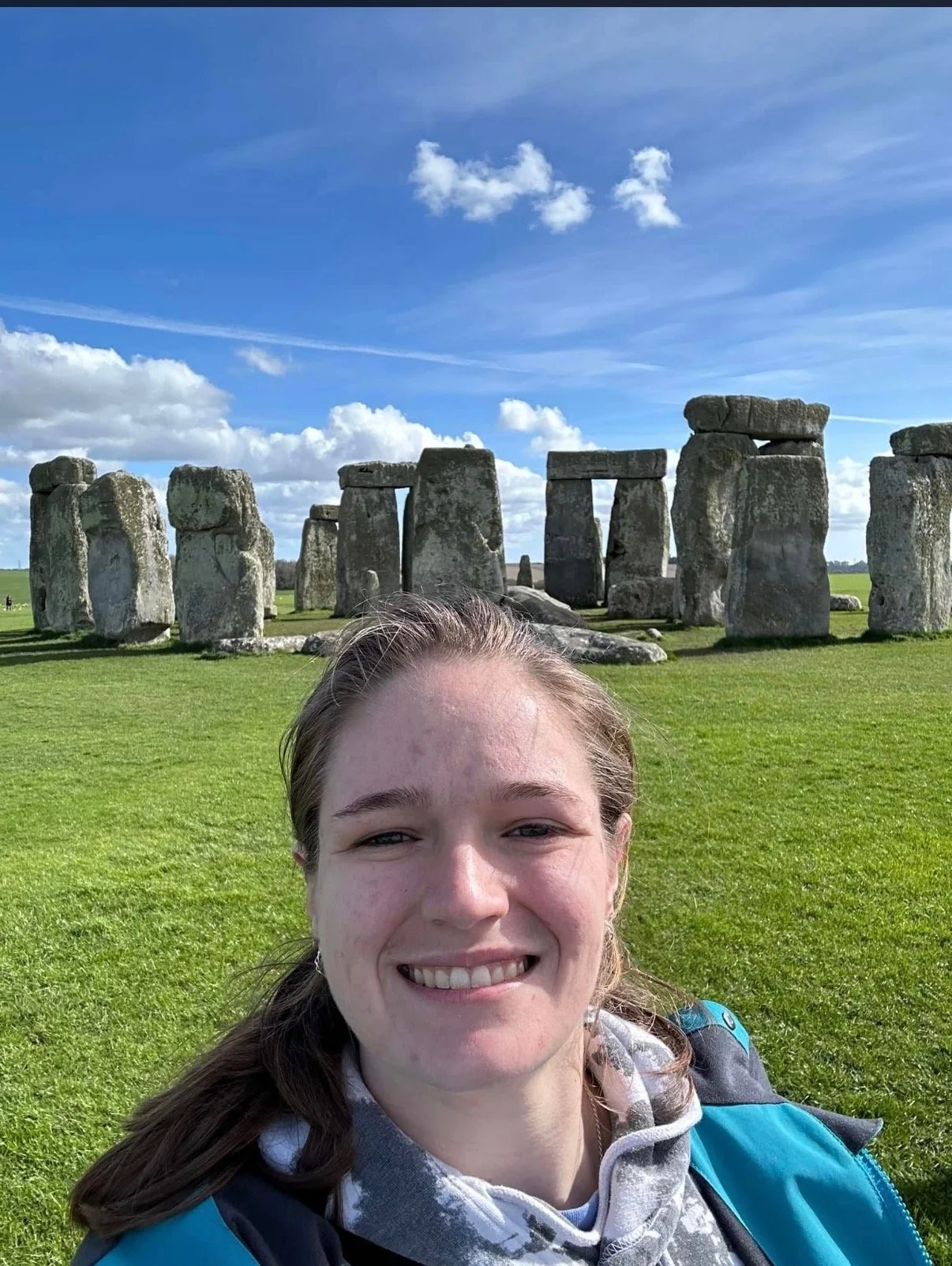 A young woman taking a selfie in front of Stonehenge on a sunny day with a blue sky and white clouds.