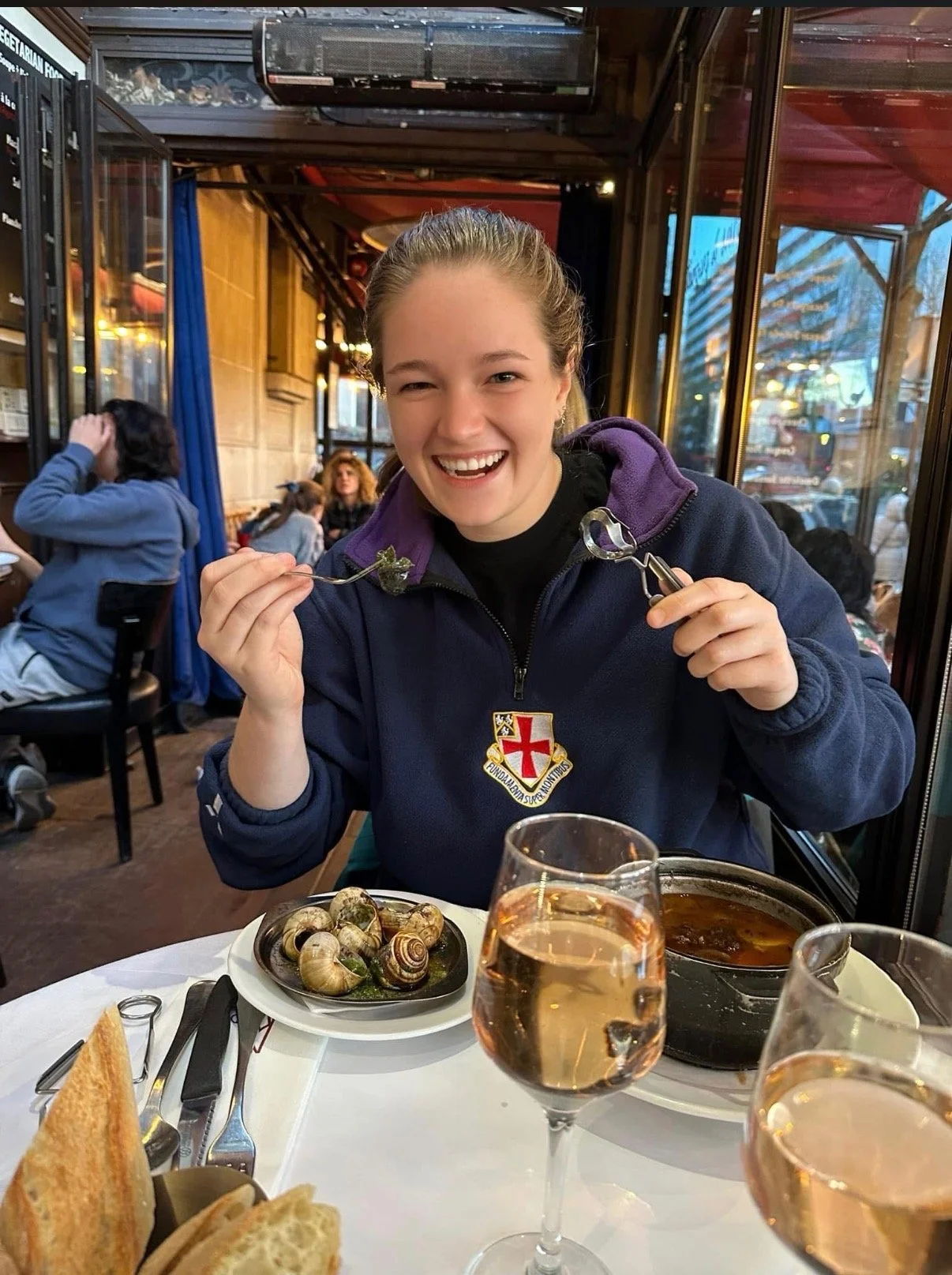 A young woman with a broad smile, wearing a navy blue jacket with a red and white cross crest, enjoys a meal at a restaurant. She holds a fork with a piece of food, and there are glasses of rosé wine and a plate of snails on the table. The restaurant has a cozy ambiance with several other diners in the background.