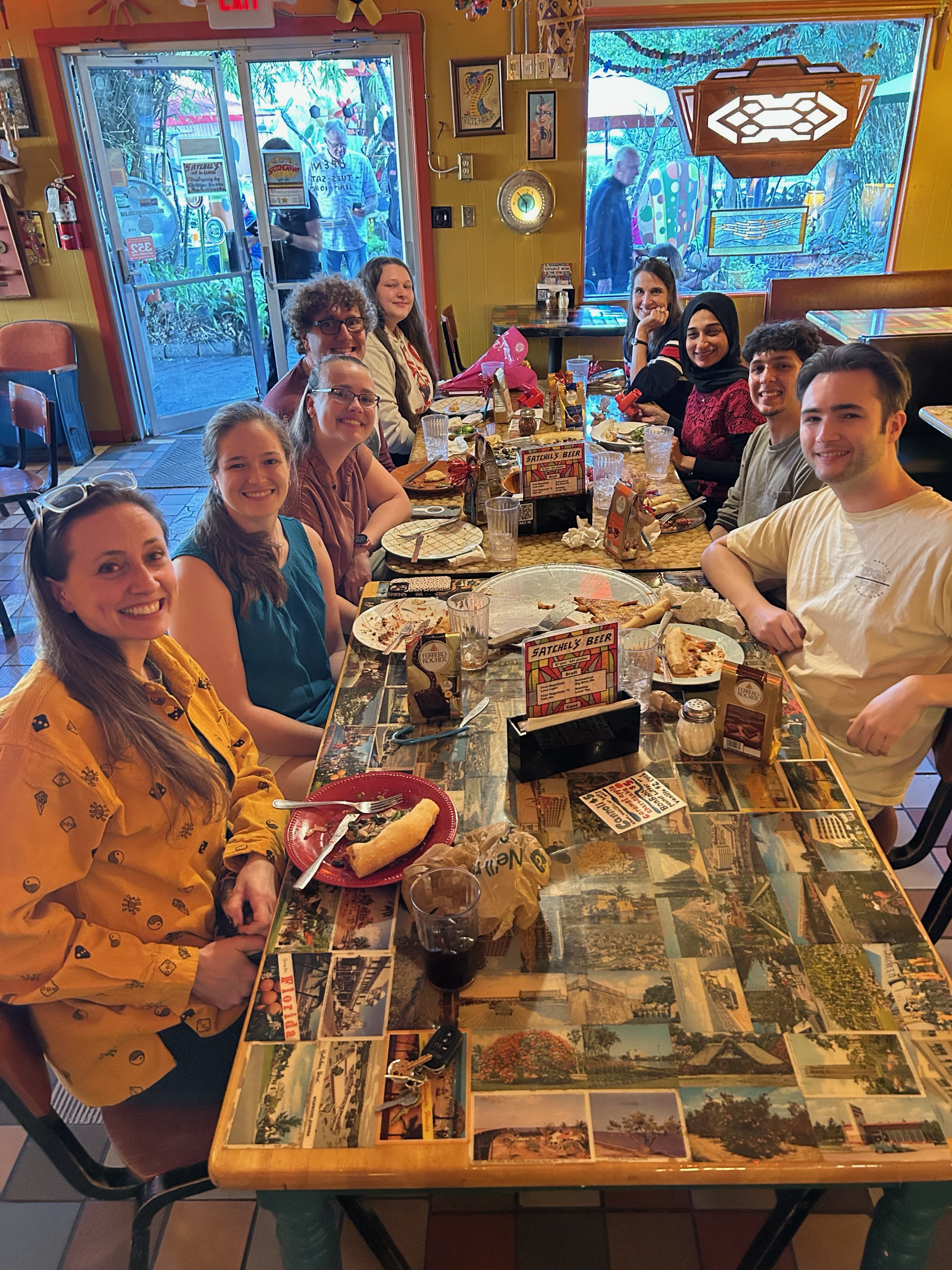 A group of nine people sitting around a long rectangular table in a restaurant, with food, drinks, and condiments on the table, smiling and looking at the camera. Outside the restaurant window, there are people visible through glass, and the interior decor includes colorful wall art and photographs of scenery.