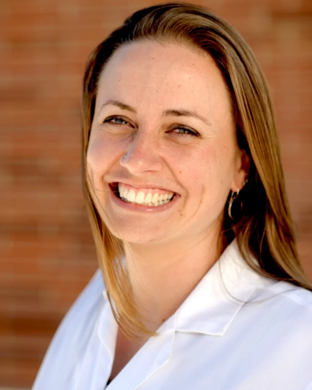 A woman with light skin, long brown hair, and a nose piercing, smiling outdoors against a blurred brick wall background.