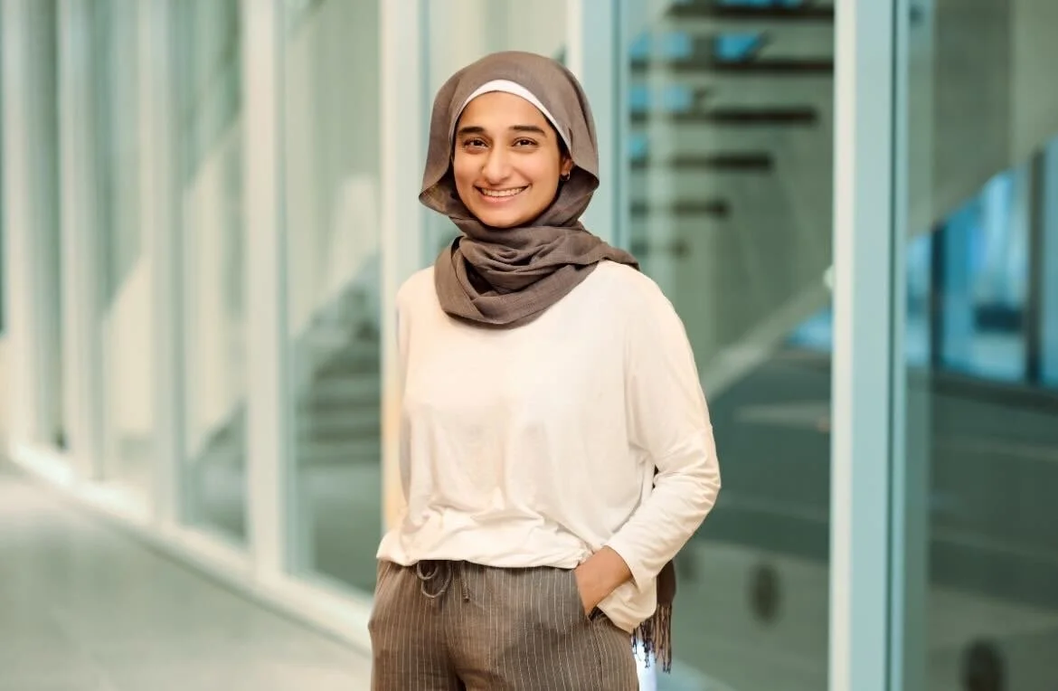 Smiling woman wearing a beige hijab, white long-sleeve top, and striped pants standing in a modern glass corridor.