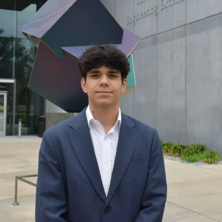 Young man in a suit standing outside a modern building with a large abstract sculpture behind him.