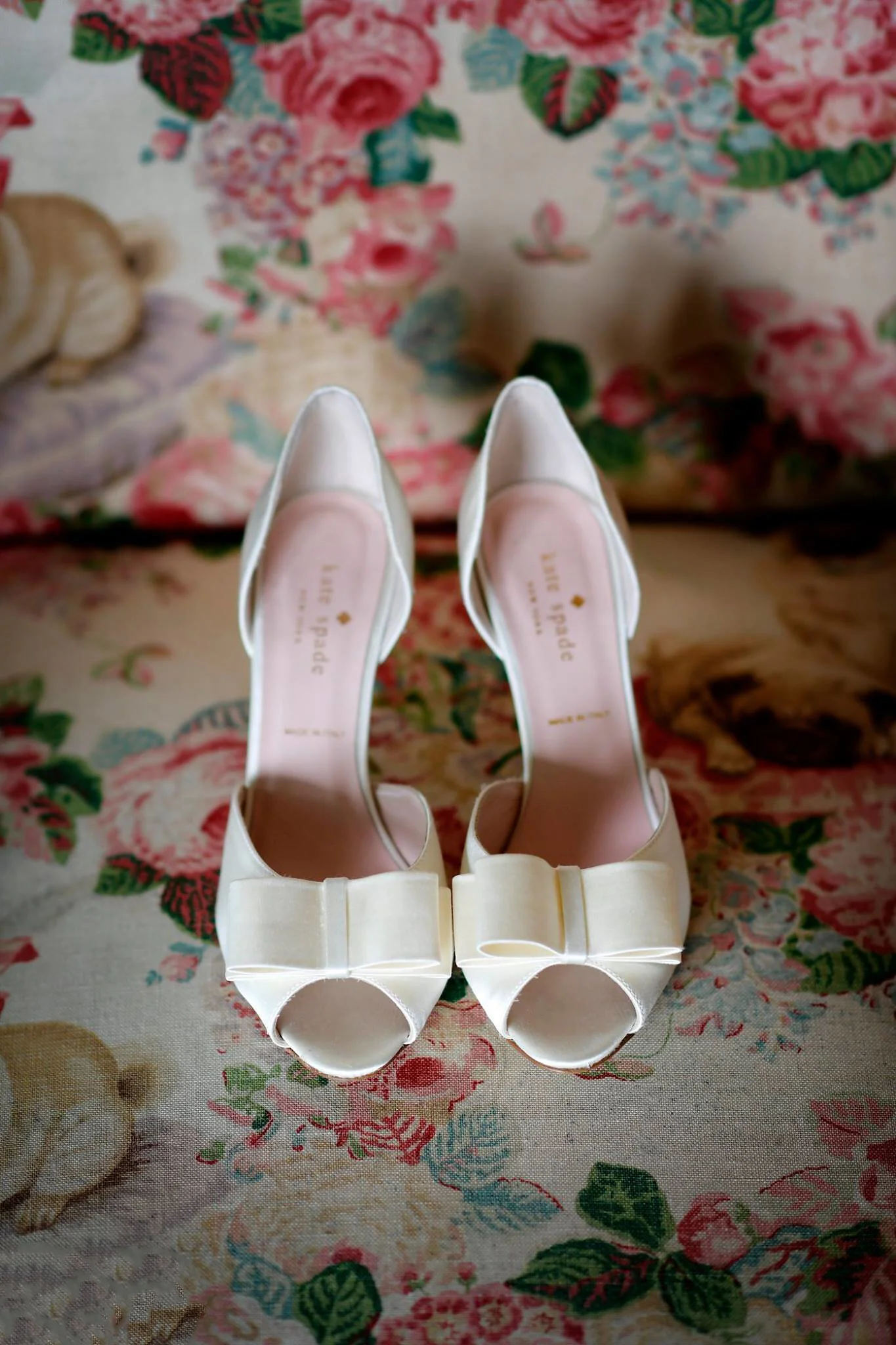 A pair of white satin high-heeled shoes with bows on the front, placed on a floral fabric surface.