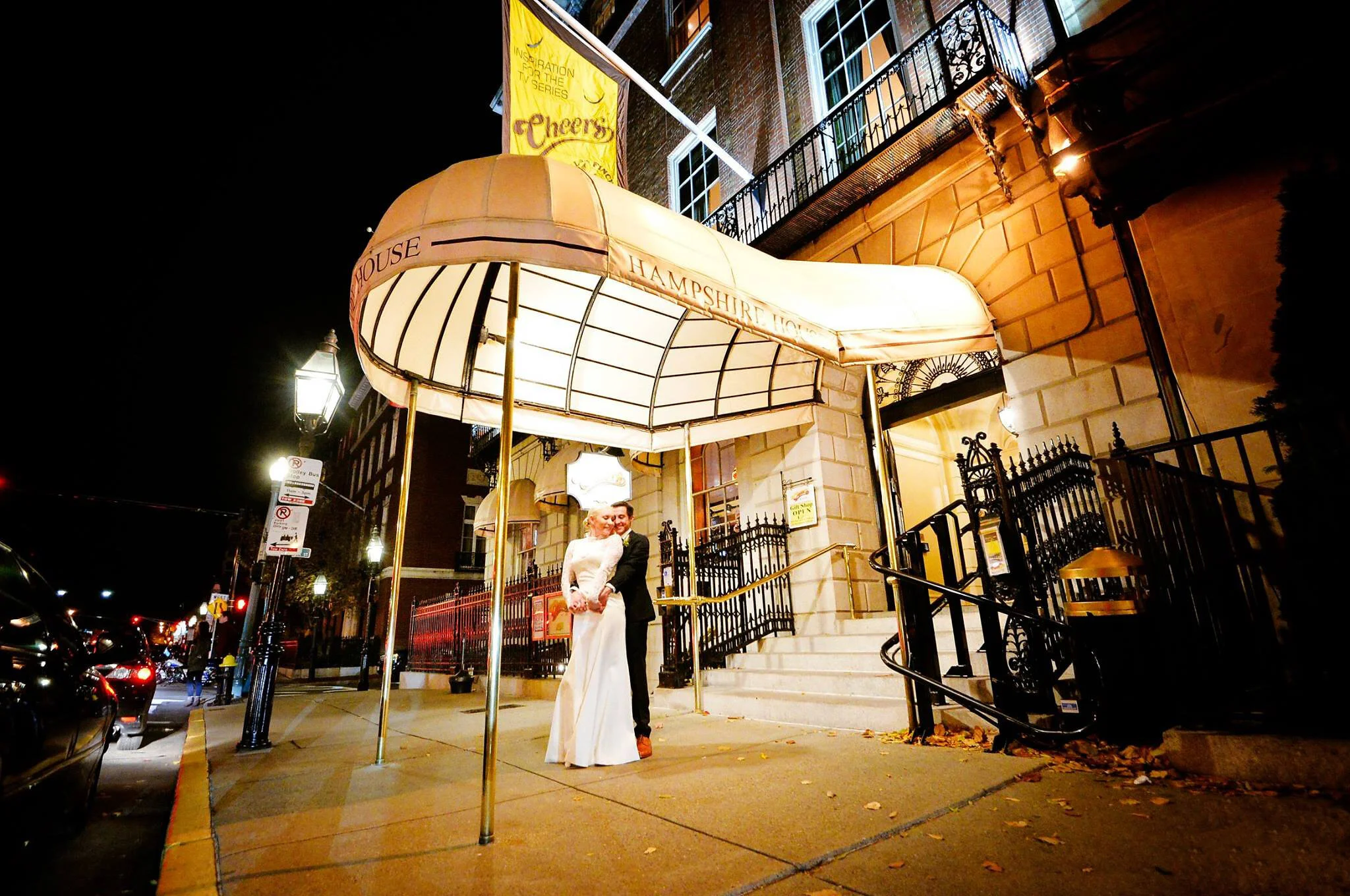 A newly married couple, dressed in white wedding attire, standing close together outside the Hampshire House on a city street at night, under a large illuminated canopy. The scene is lit by street lamps, with cars and pedestrians visible in the backg
