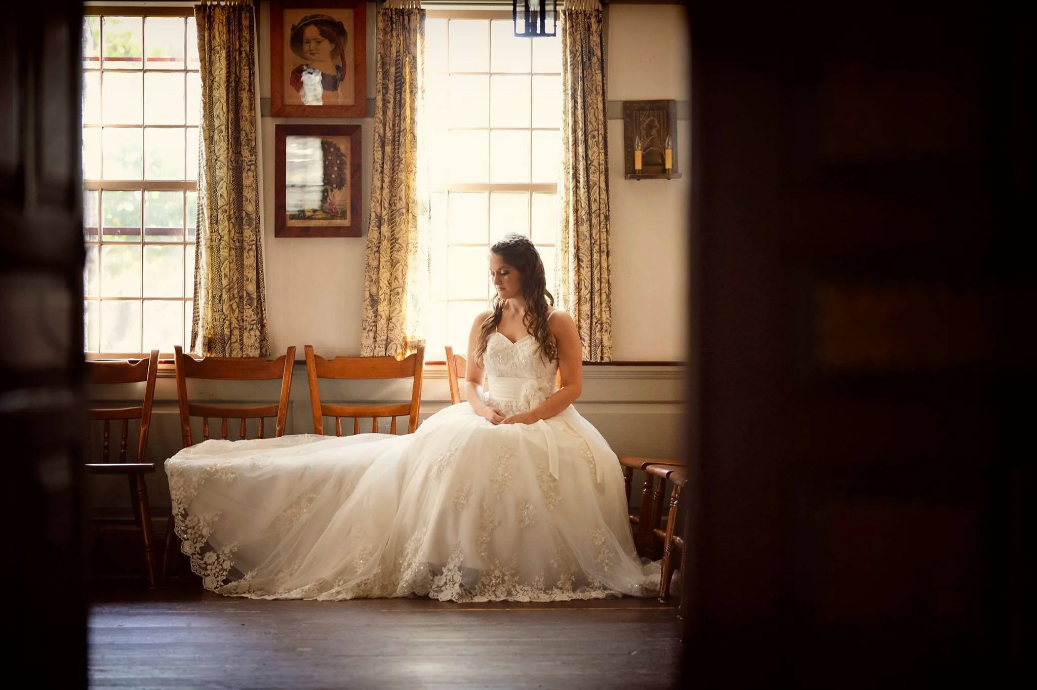 A bride in a white wedding dress sitting on a wooden bench in a warmly lit room with large windows and floral curtains.