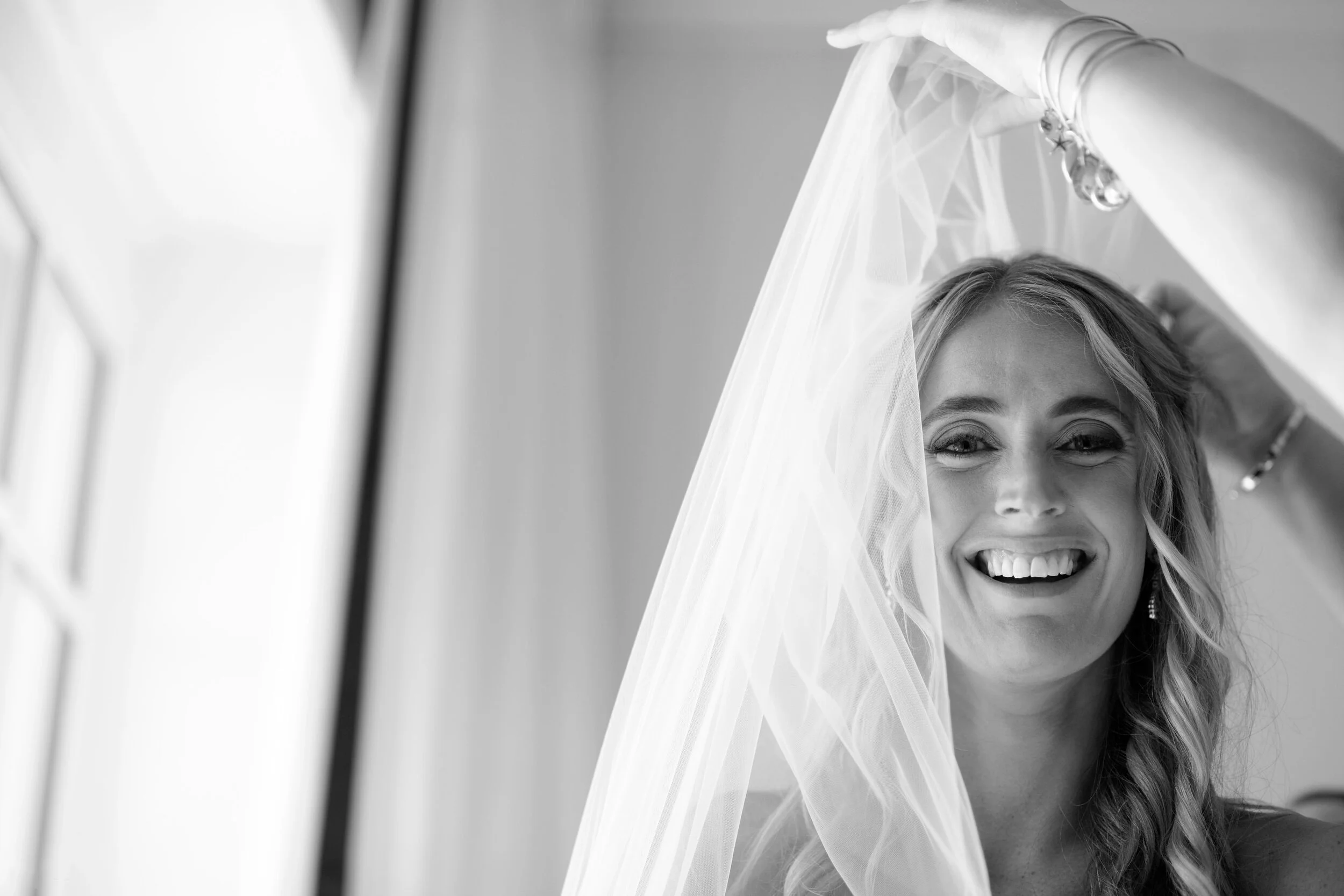 A smiling woman with a veil over her head, adjusting it with her hand, illuminated by natural light from a nearby window.
