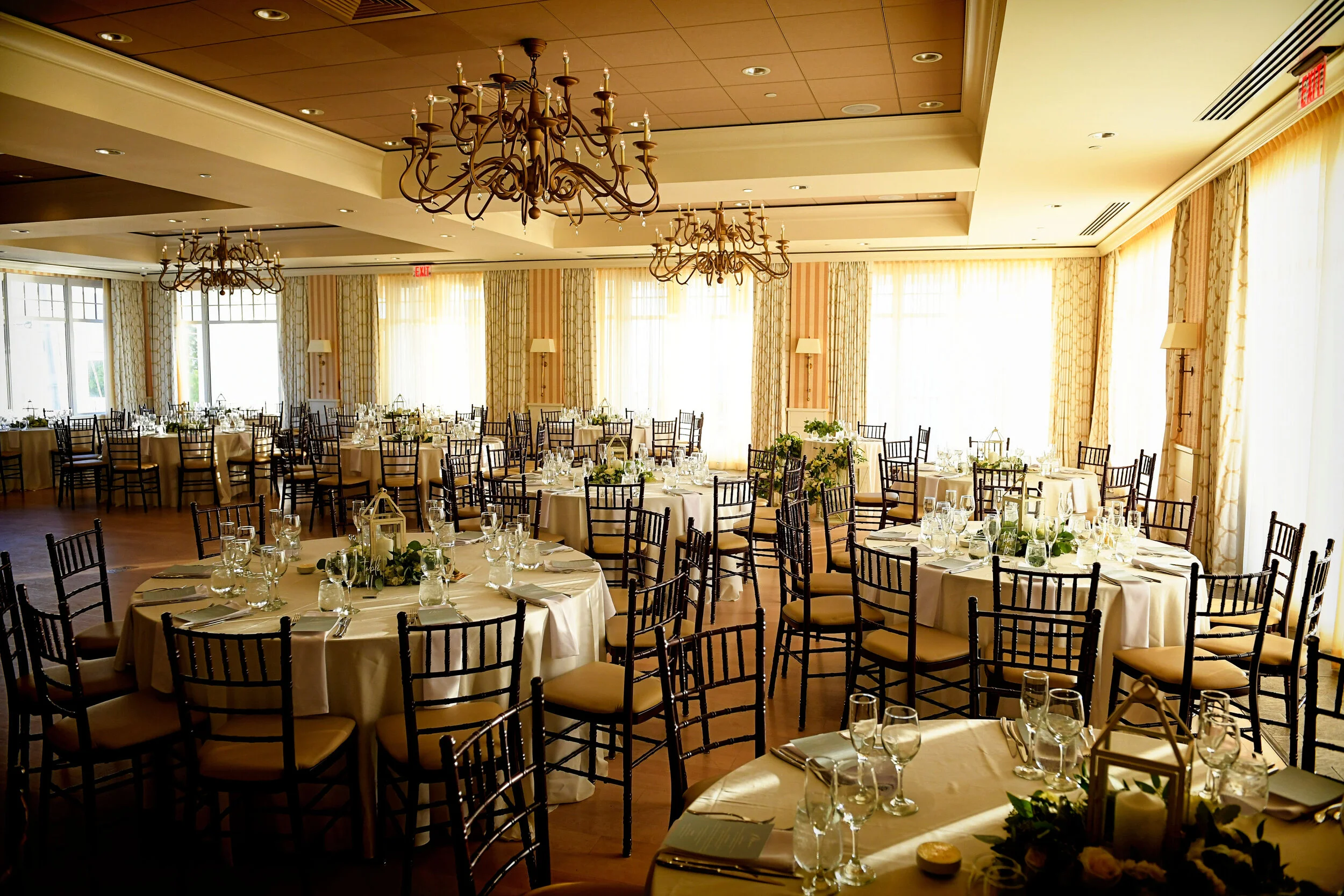Elegant banquet hall with round tables covered in white tablecloths, set with glassware and napkins, decorated with candles and greenery, illuminated by natural light from large windows, and chandeliers hanging from the ceiling.