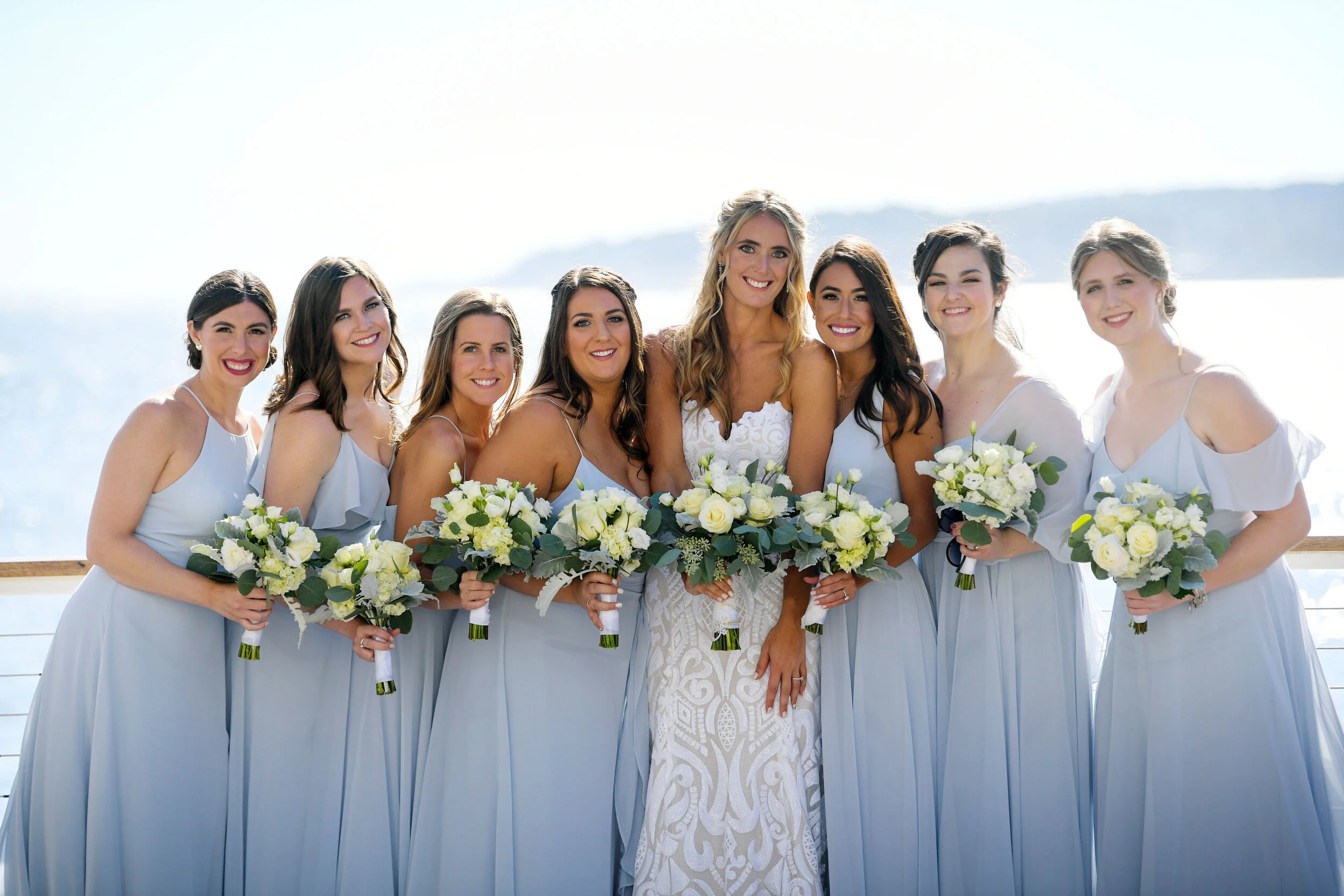 Group of nine women, including bride and bridesmaids, dressed in light blue and white, holding bouquets of white and green flowers, standing outdoors near water with hills in background.