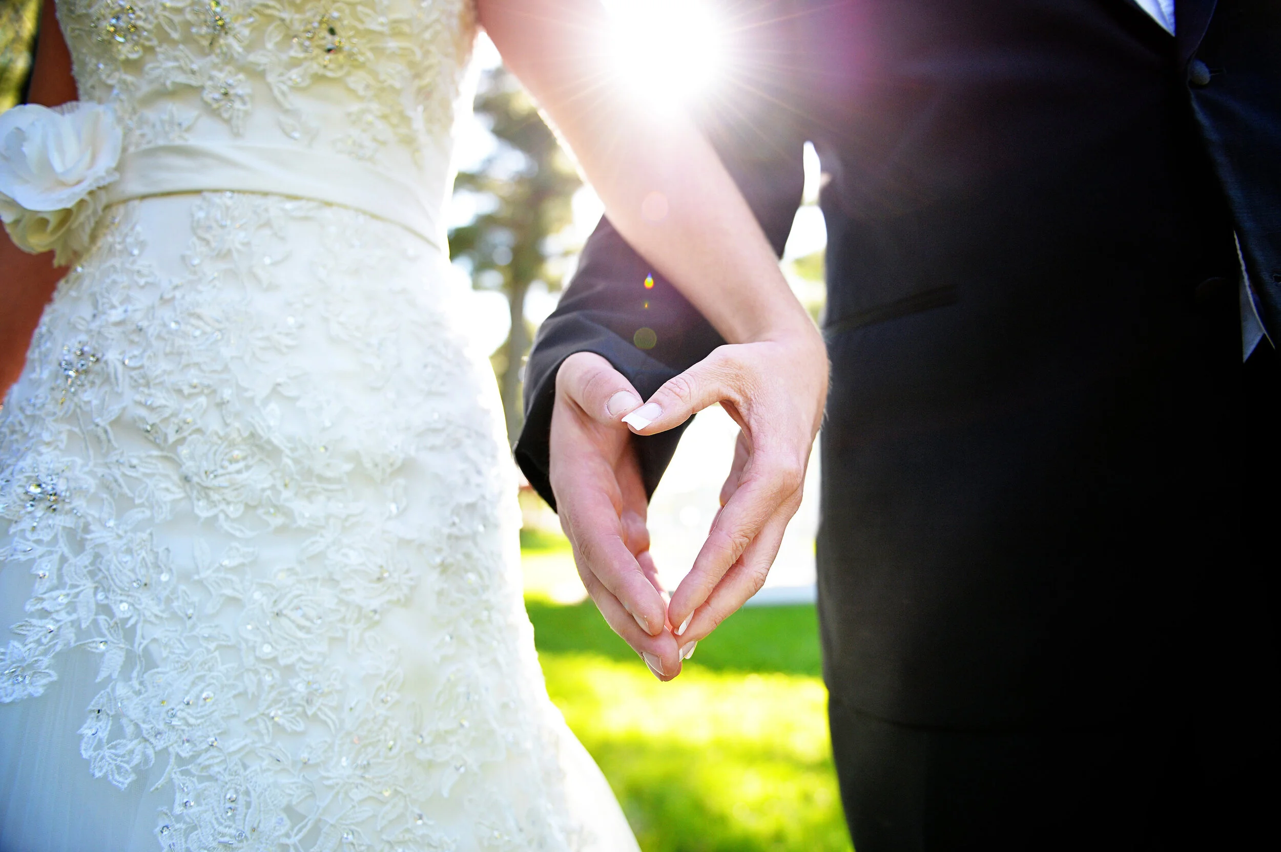 Close-up of a bride and groom holding hands outdoors during daytime, with sunlight shining behind them. The bride is wearing a white wedding dress with lace and embellishments, and the groom is wearing a dark suit.