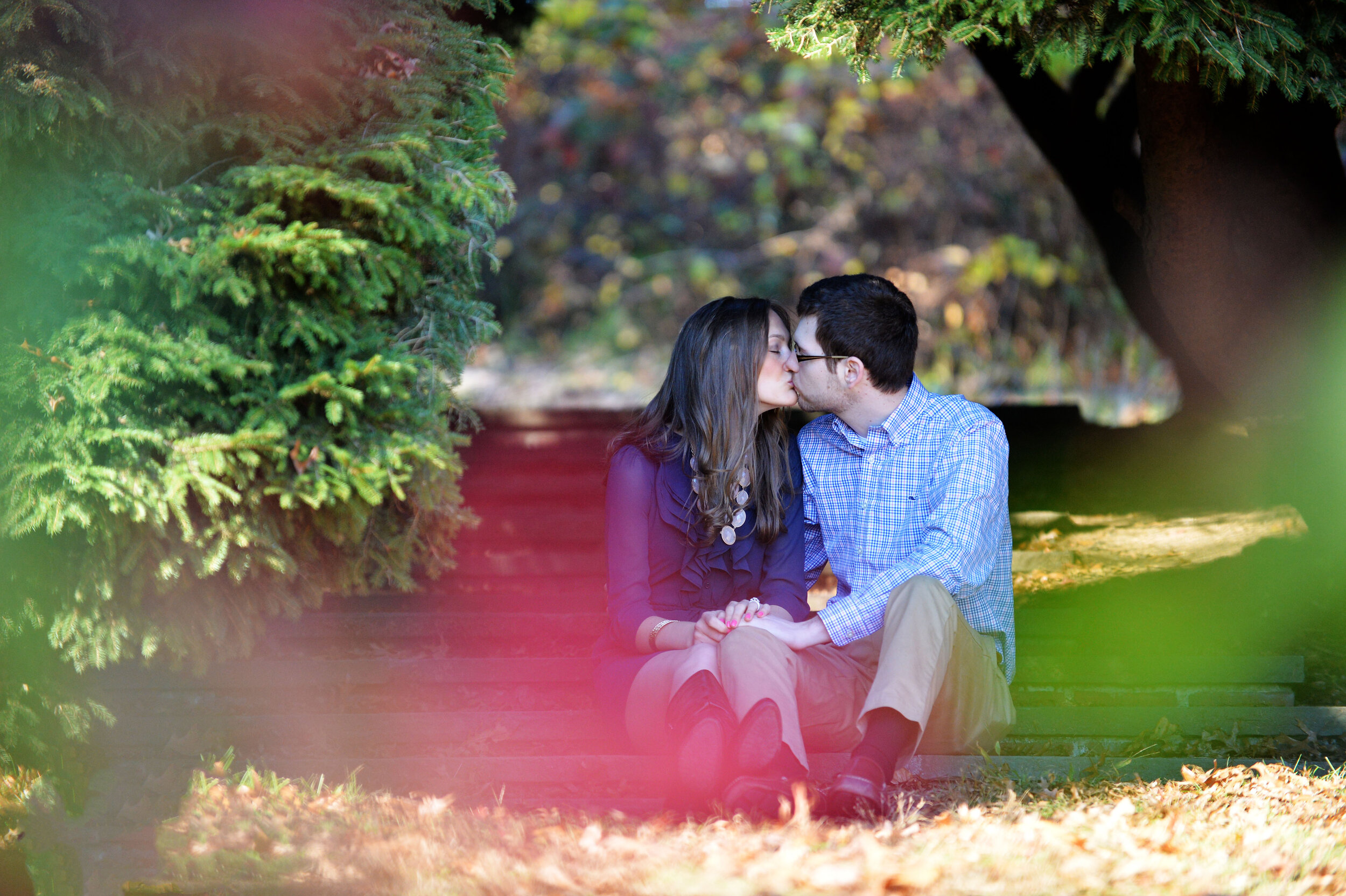 A young couple sitting on wooden steps outdoors, kissing with trees and autumn foliage in the background.