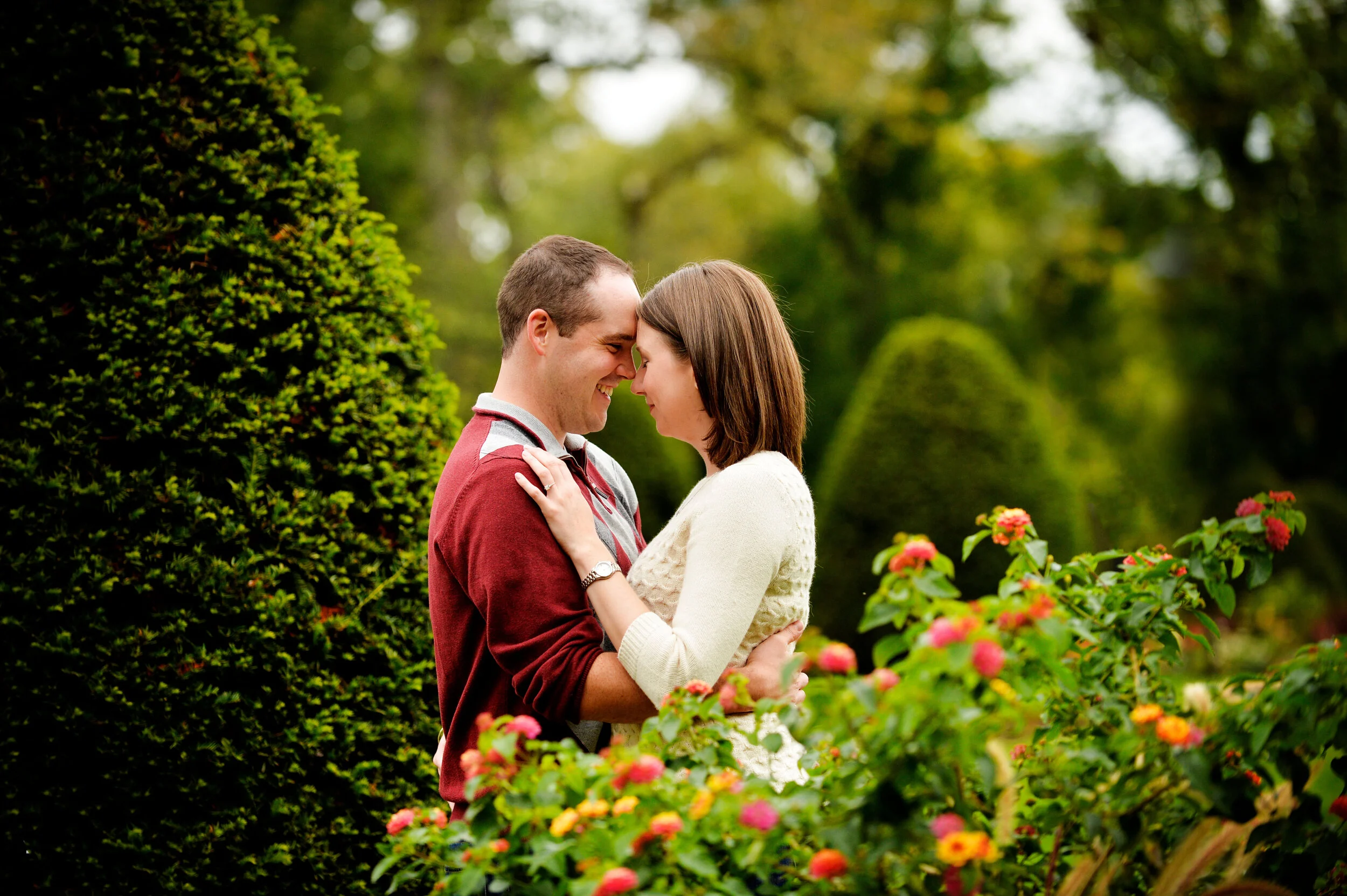 A couple stands close together in a garden, touching foreheads and smiling with their eyes closed, surrounded by green bushes and colorful flowers.