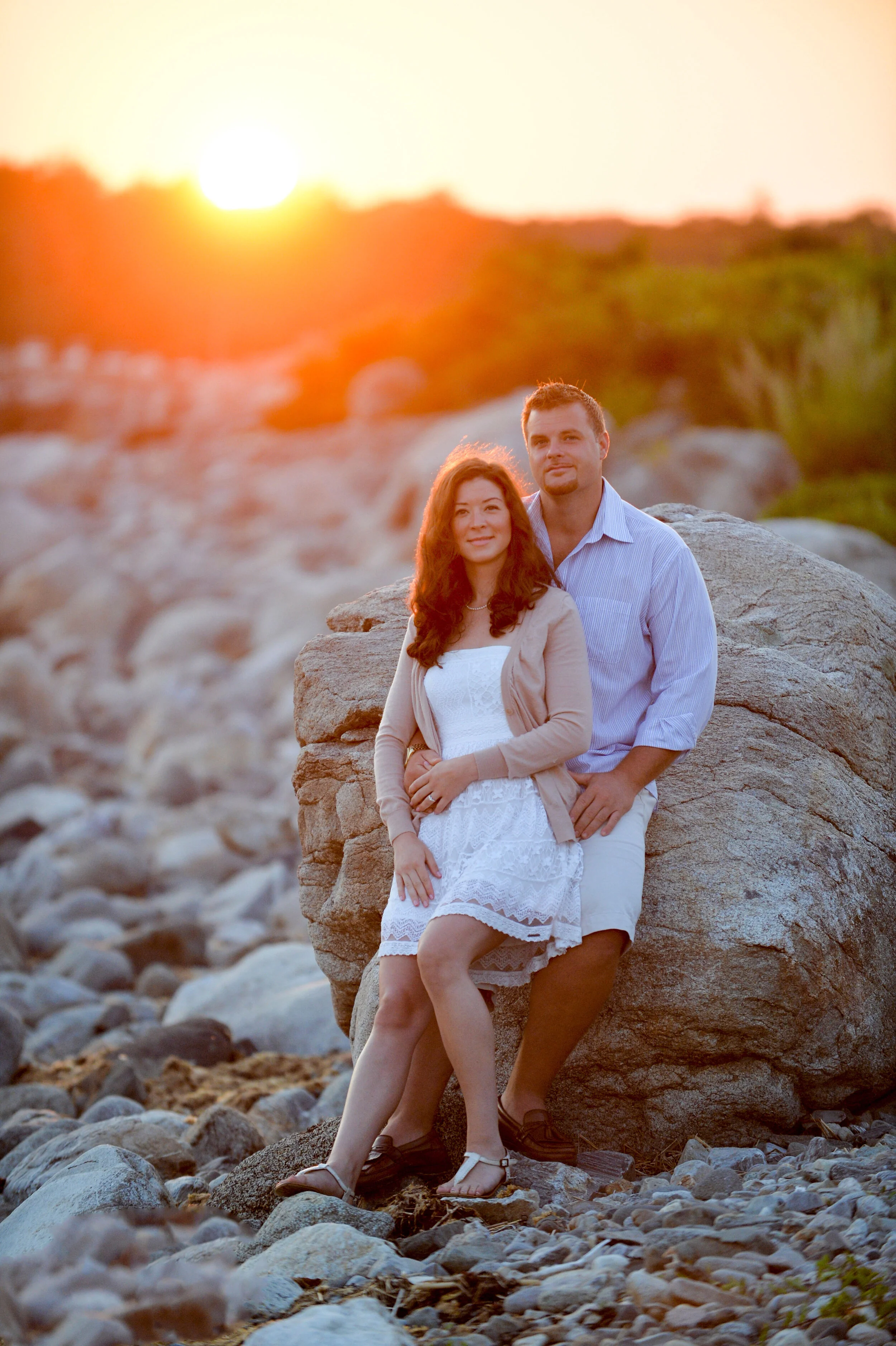 A couple sitting on rocks at sunset, with a rocky beach and distant trees in the background.