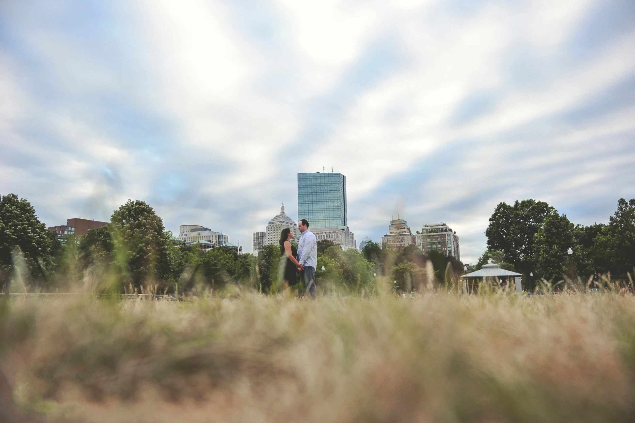 A couple holding hands in a park with a city skyline in the background, including tall buildings and a cloudy sky.