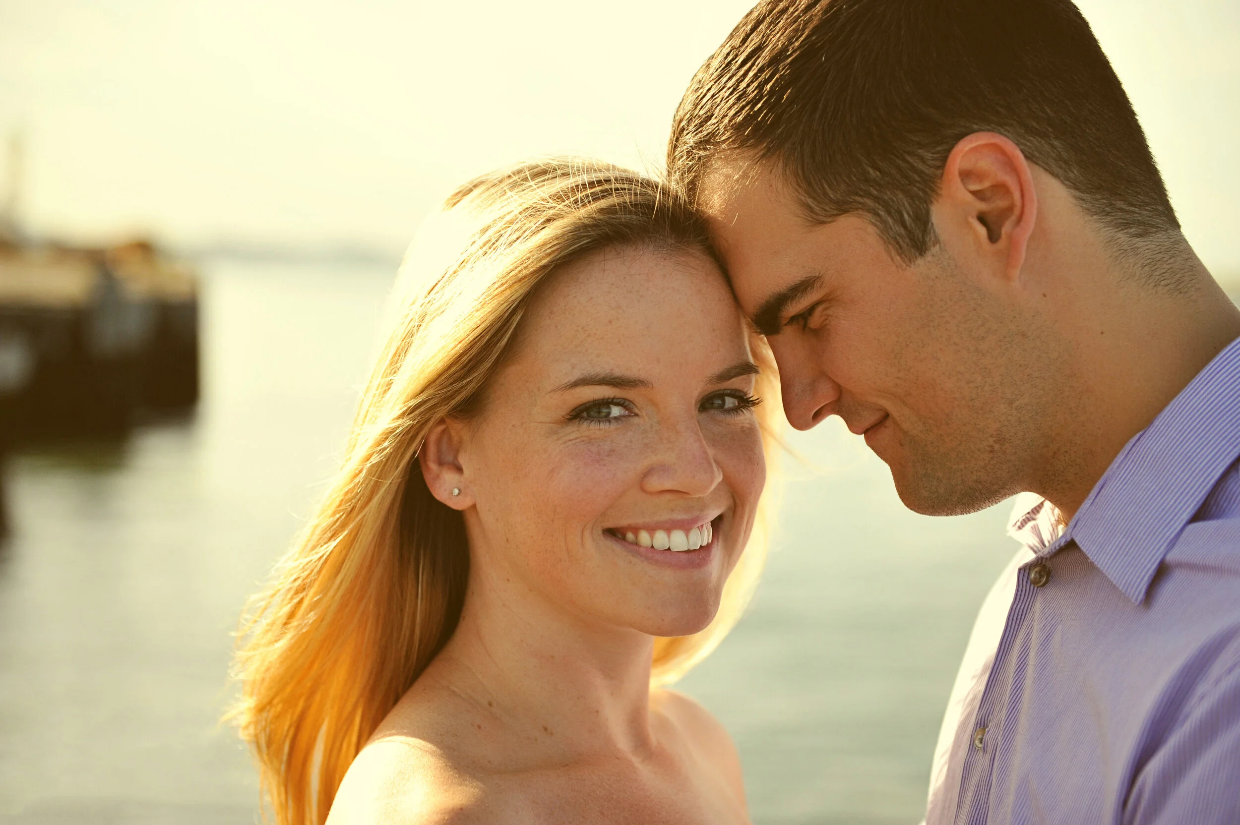 A smiling woman and a man with their foreheads touching outdoors near water at sunset.