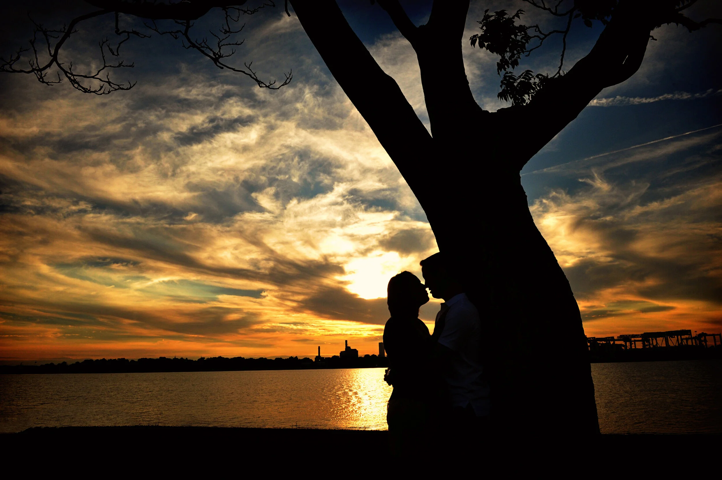 Silhouette of a couple kissing next to a large tree by water during a colorful sunset with clouds and city skyline in the background.