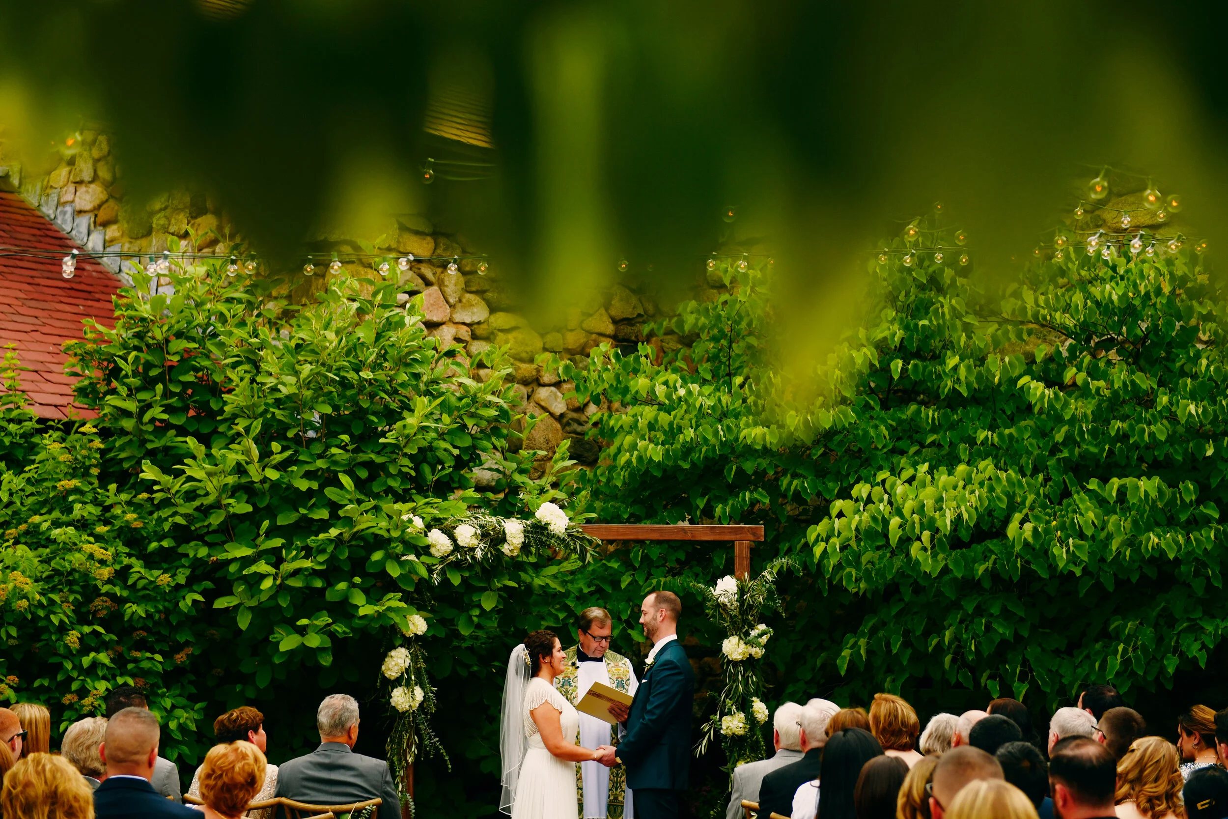 Outdoor wedding ceremony with a couple exchanging vows under a floral arch, surrounded by greenery and guests seated in rows.