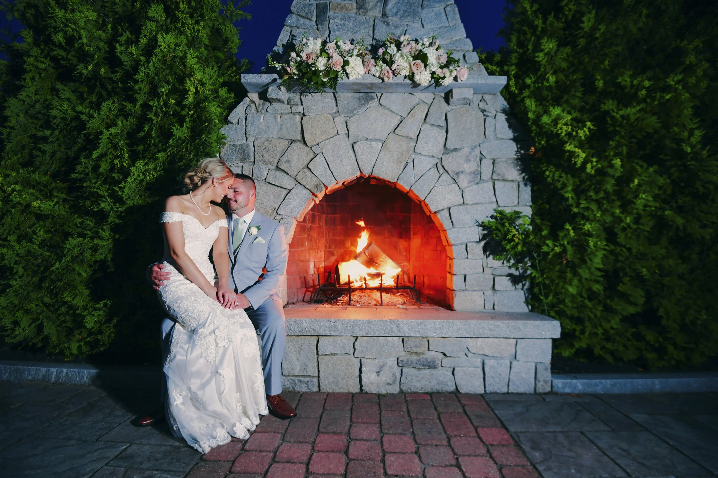 Bride and groom sitting by a lit stone fireplace with floral decoration.