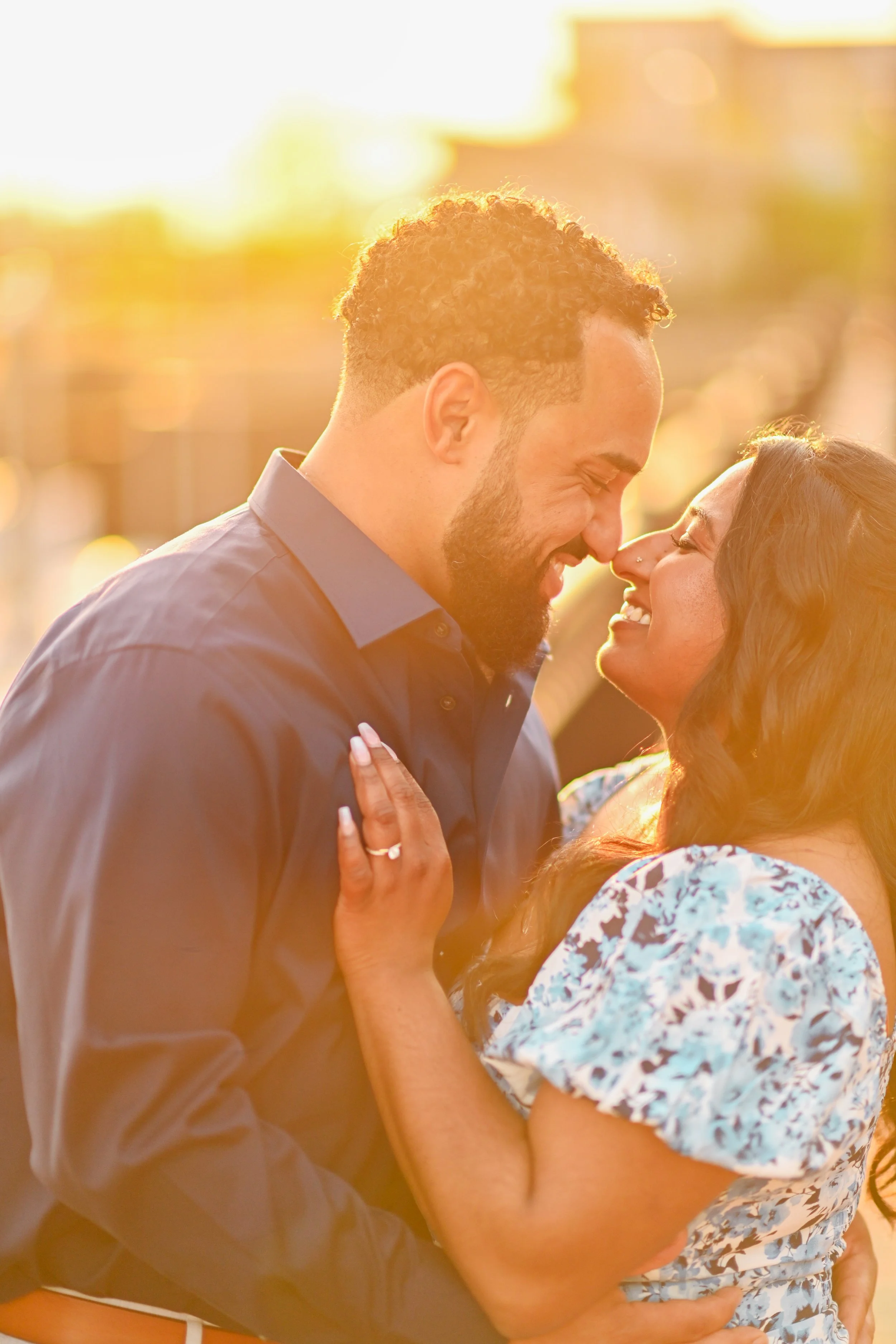 A couple smiling and embracing each other outdoors during sunset, their faces close and noses touching.