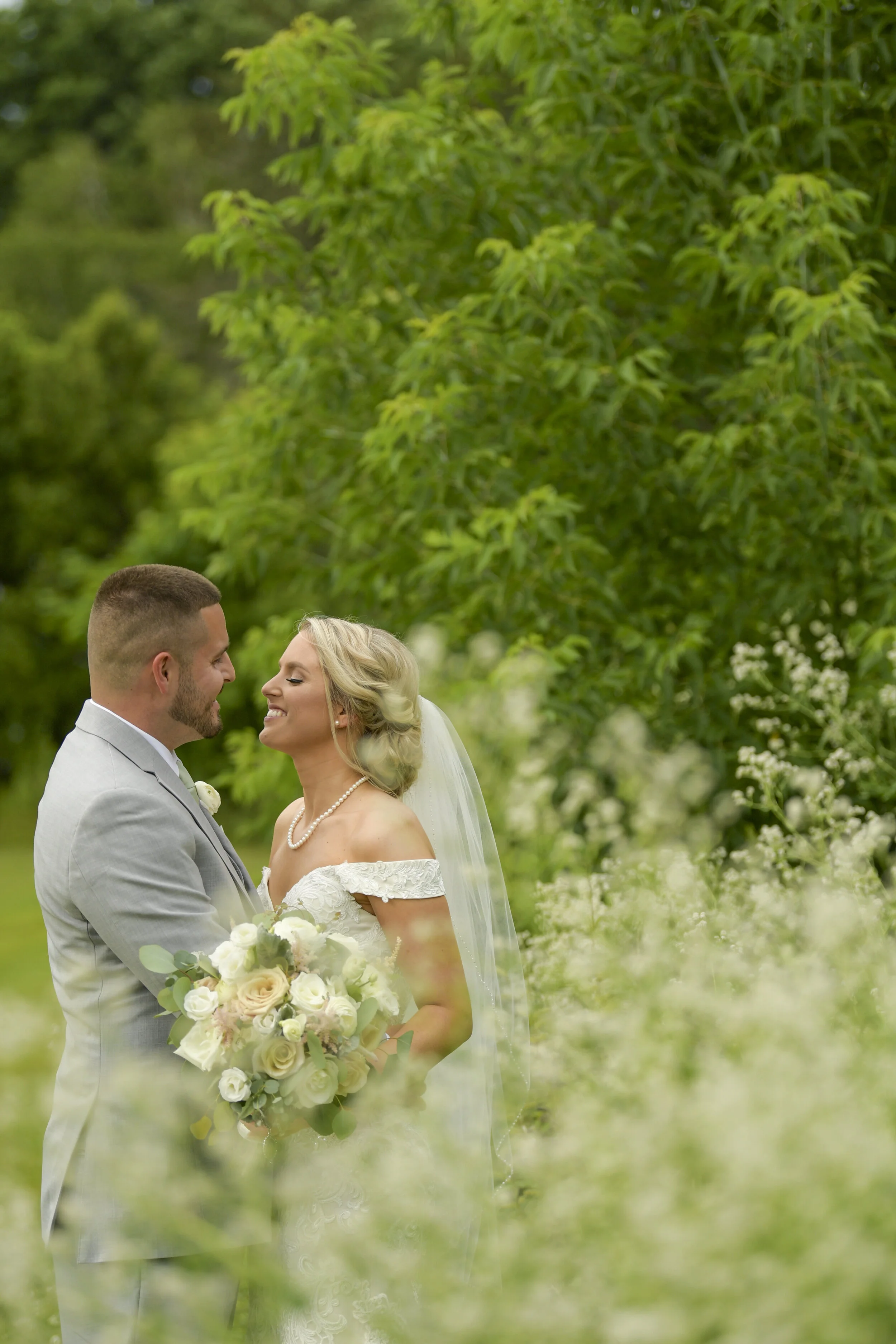 A bride and groom smiling at each other amidst lush green foliage, with the bride holding a bouquet of white flowers and wearing a veil.
