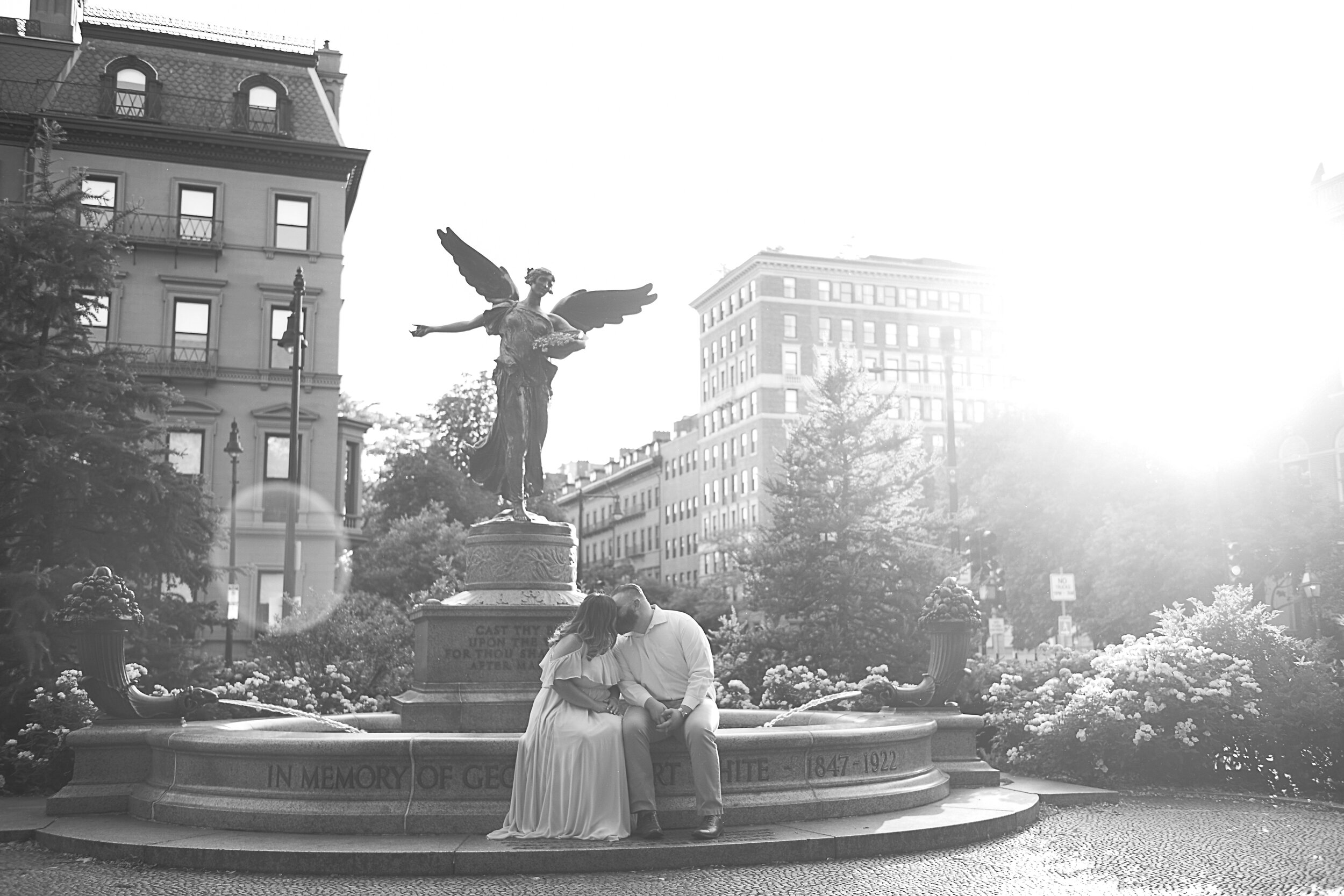 A couple sits close together on a fountain in front of a statue of an angel in an urban park, with tall buildings and trees in the background, and bright sunlight.