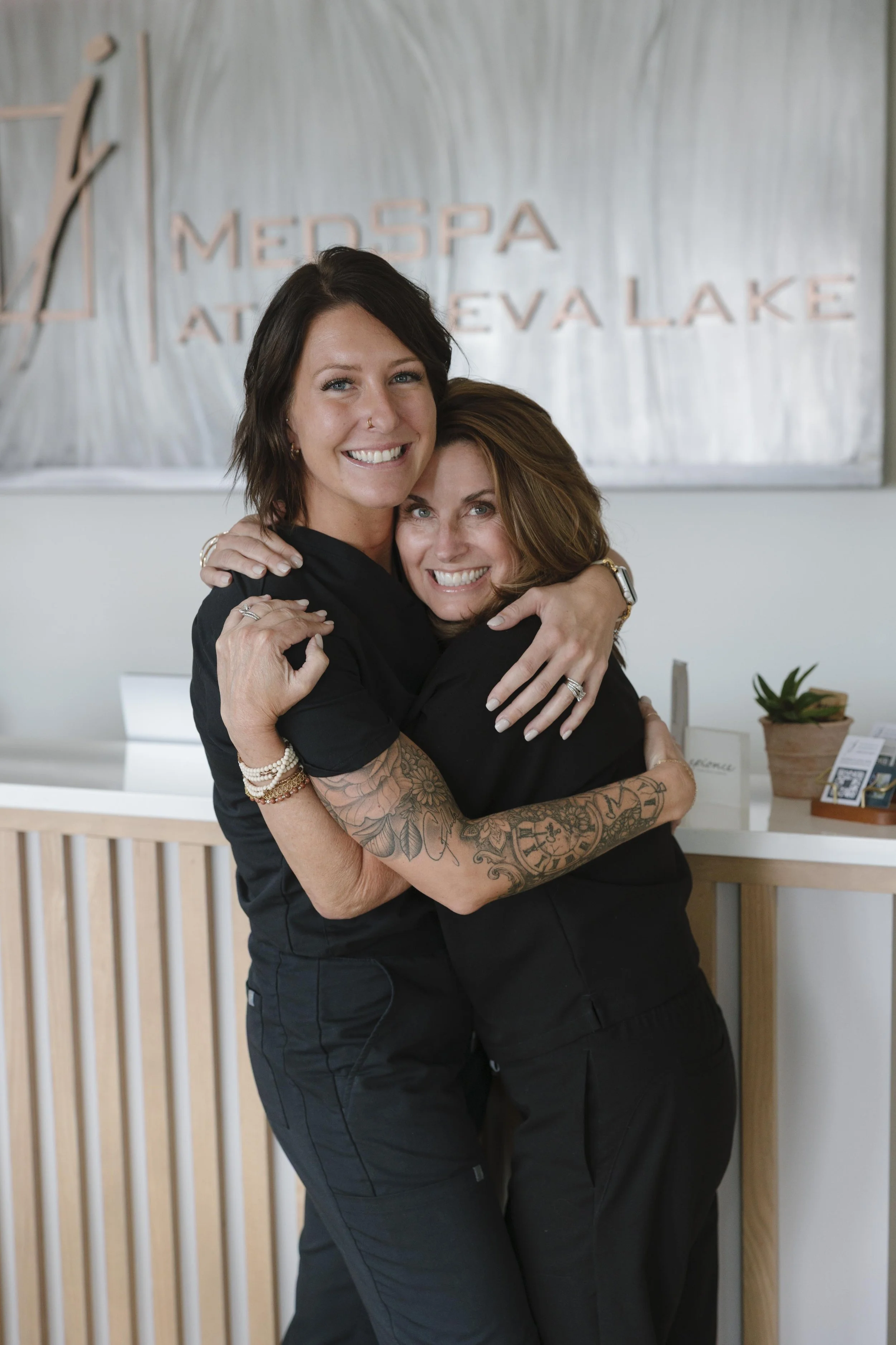 Two women in black scrubs smiling and laughing together in a medical spa reception area