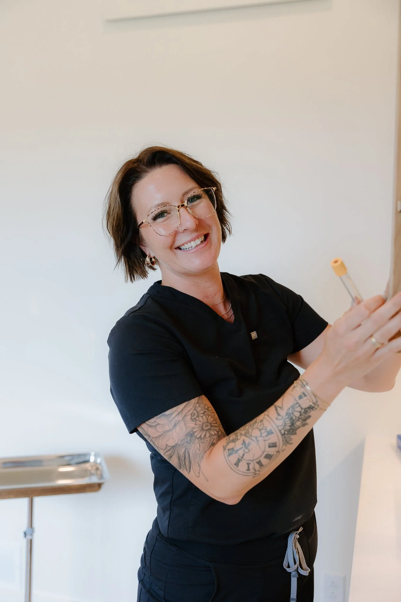 A smiling woman with glasses and short brown hair writing on a board in a medical or dental office, wearing black scrubs and tattoos on her arm.
