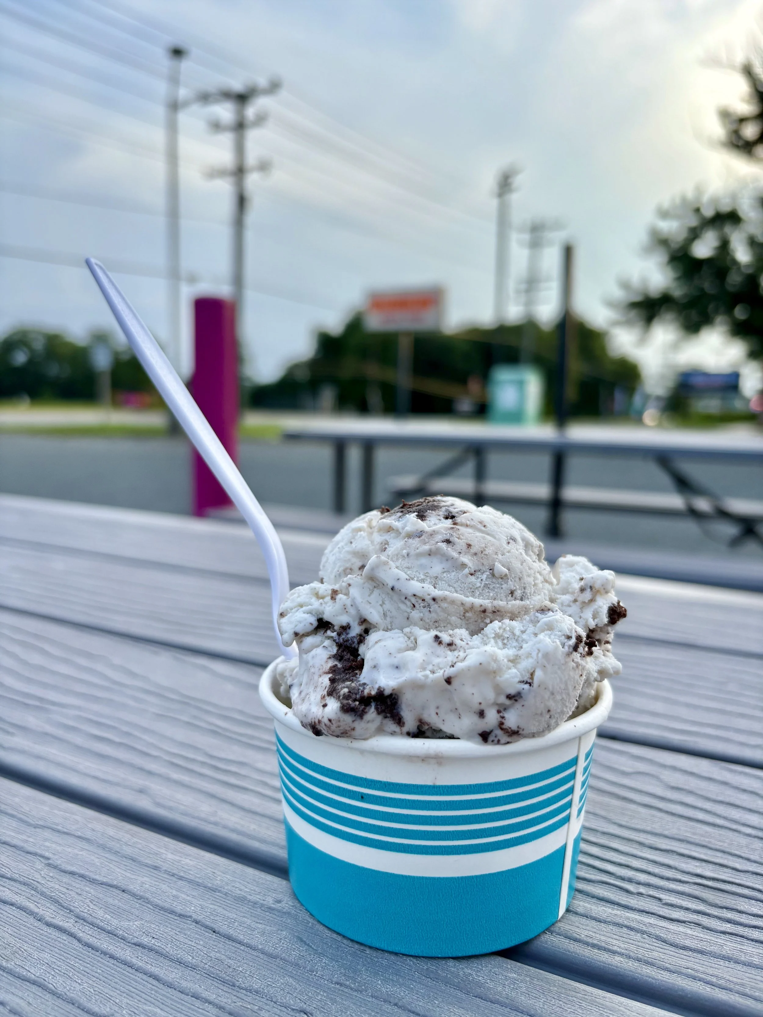 A cup of cookies and cream ice cream with a plastic spoon on a wooden outdoor table, with a blurred background of the cityscape.