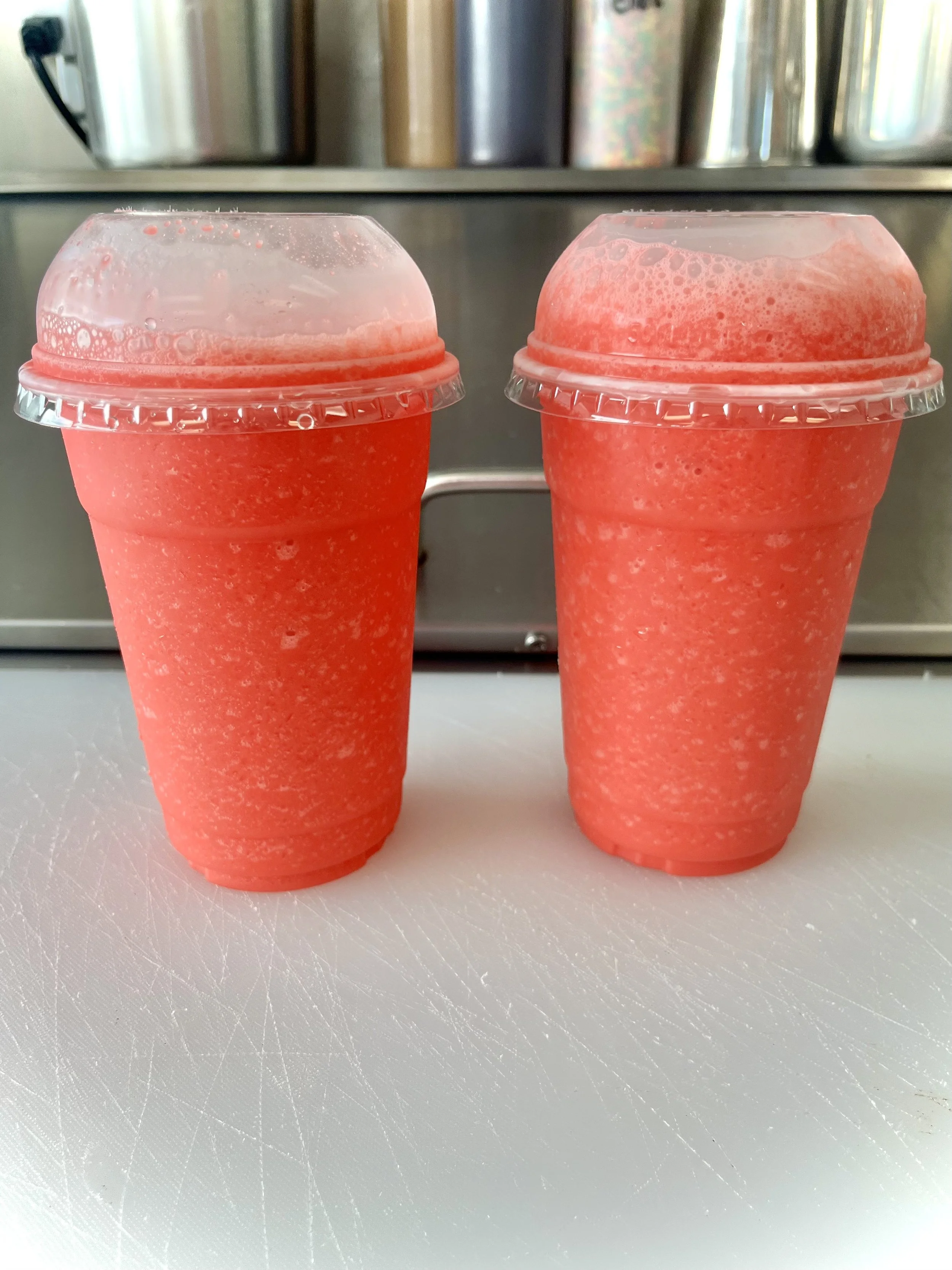 Two cups of pink slush drinks with clear dome lids on a white cutting board.