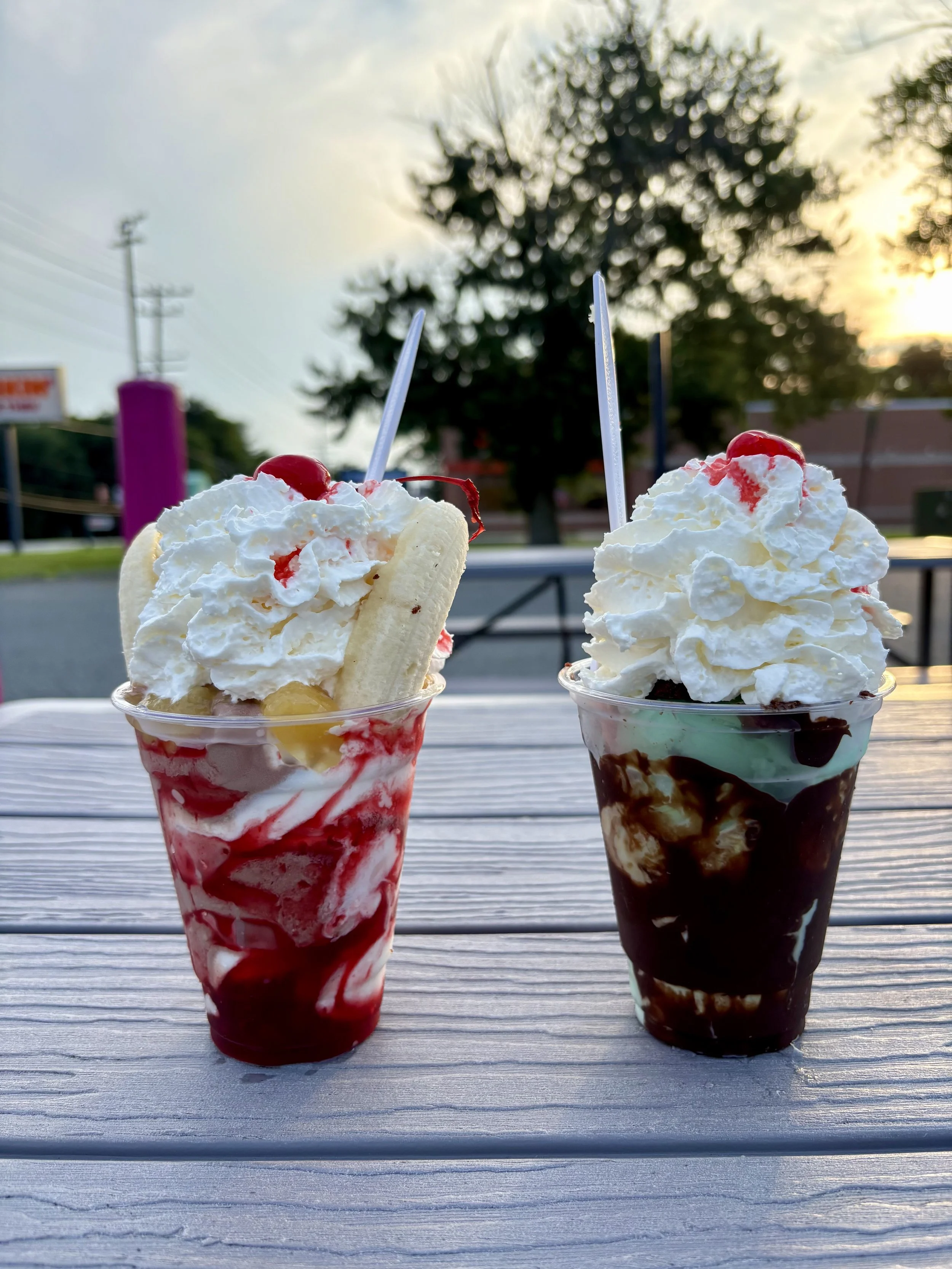 Two cups of ice cream sundaes topped with whipped cream and cherries, served on a wooden table outdoors at sunset.