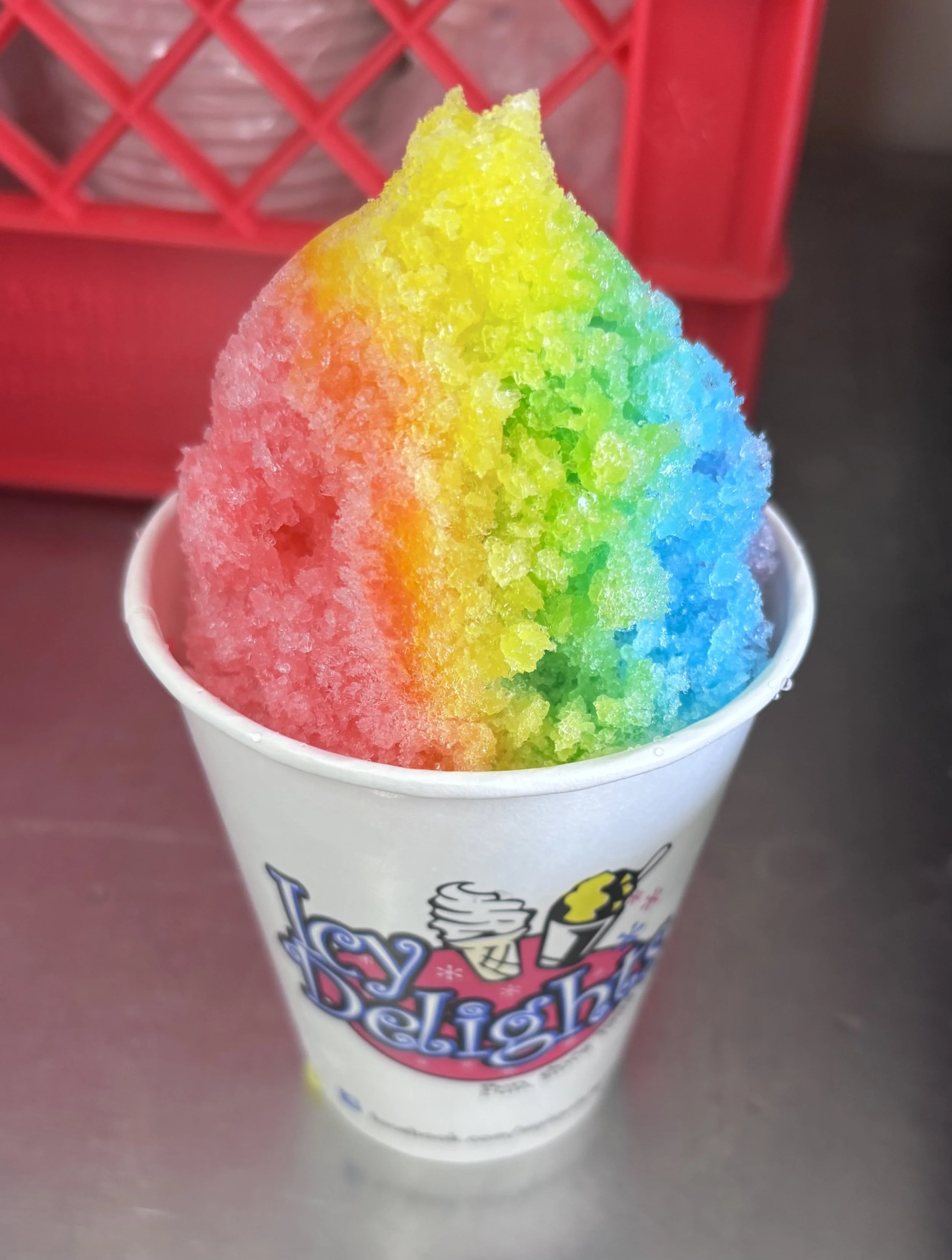 Rainbow-colored shaved ice in a white cup with a 'Icy Delights' logo.