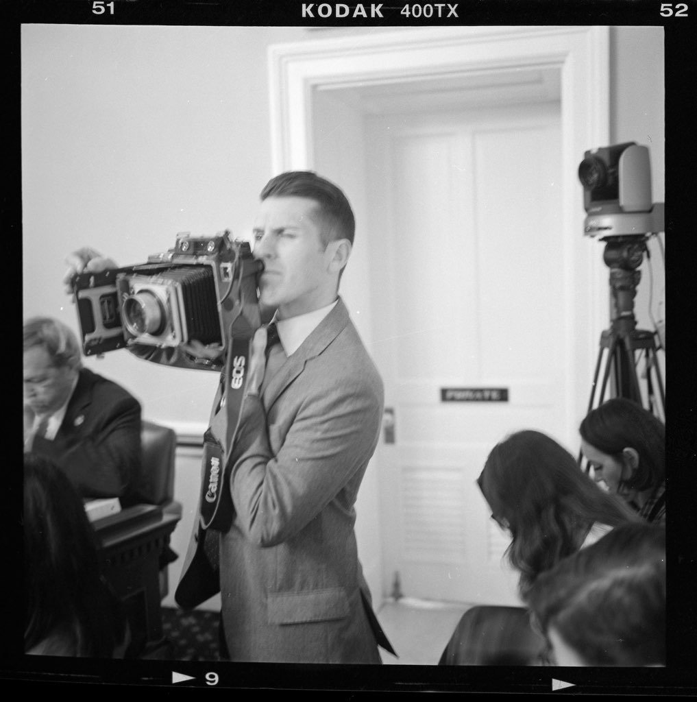 A man in a suit operating a large professional camera during a meeting or conference, with two women seated and another man visible to the side in a room with a camera on a tripod and a door in the background.