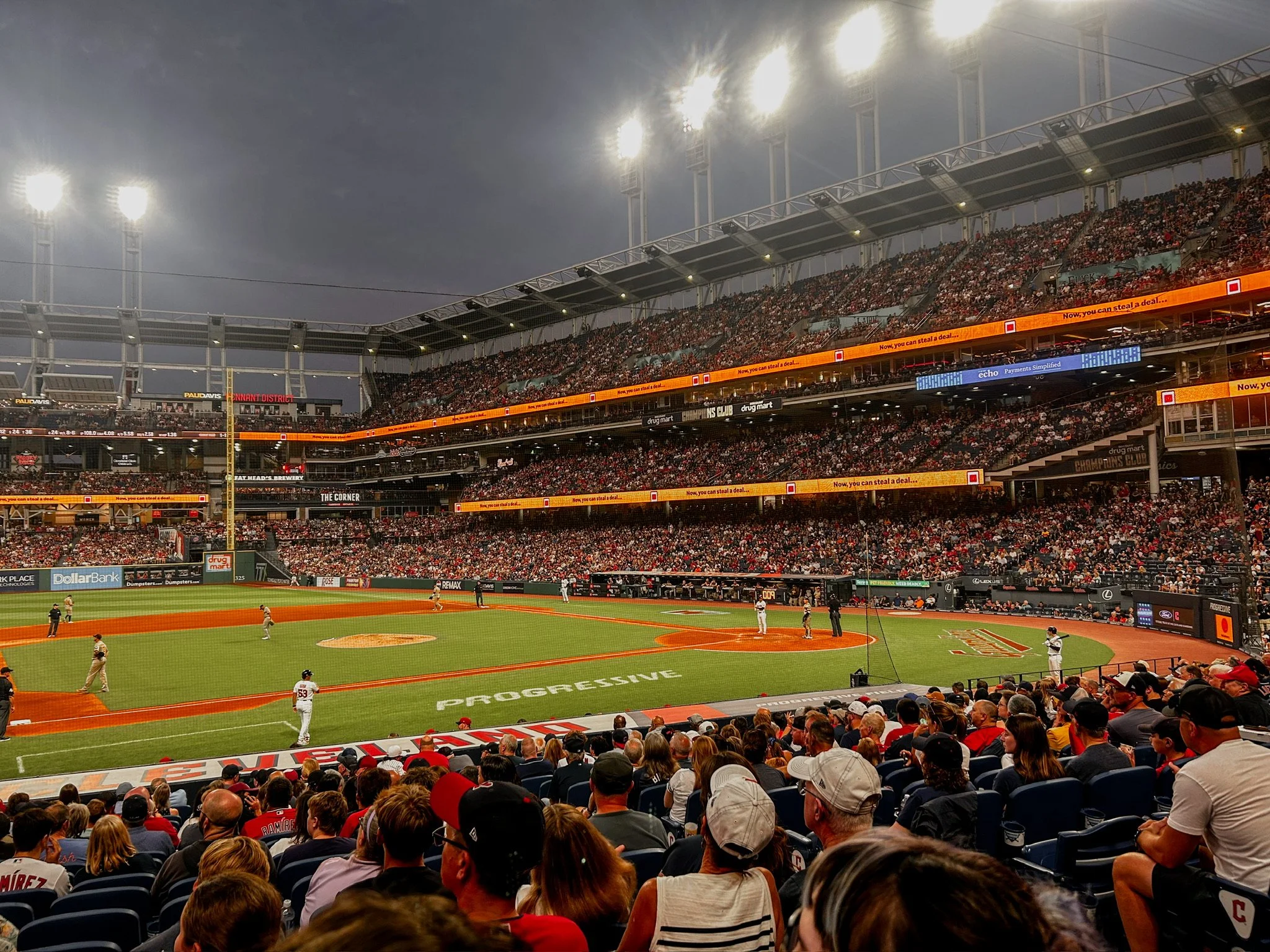 A baseball Stadium - a baseball field with players surrounded by a large 3 tier stands with bright lights