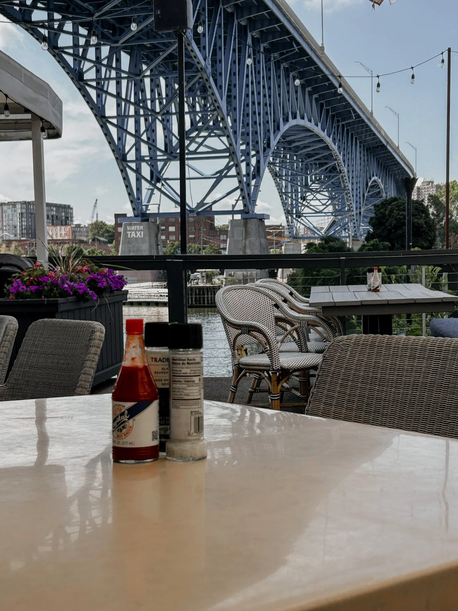 A restauarant table on a patio over looking a river with a blue bridge over the top. the table has hot sauce and salt and pepper