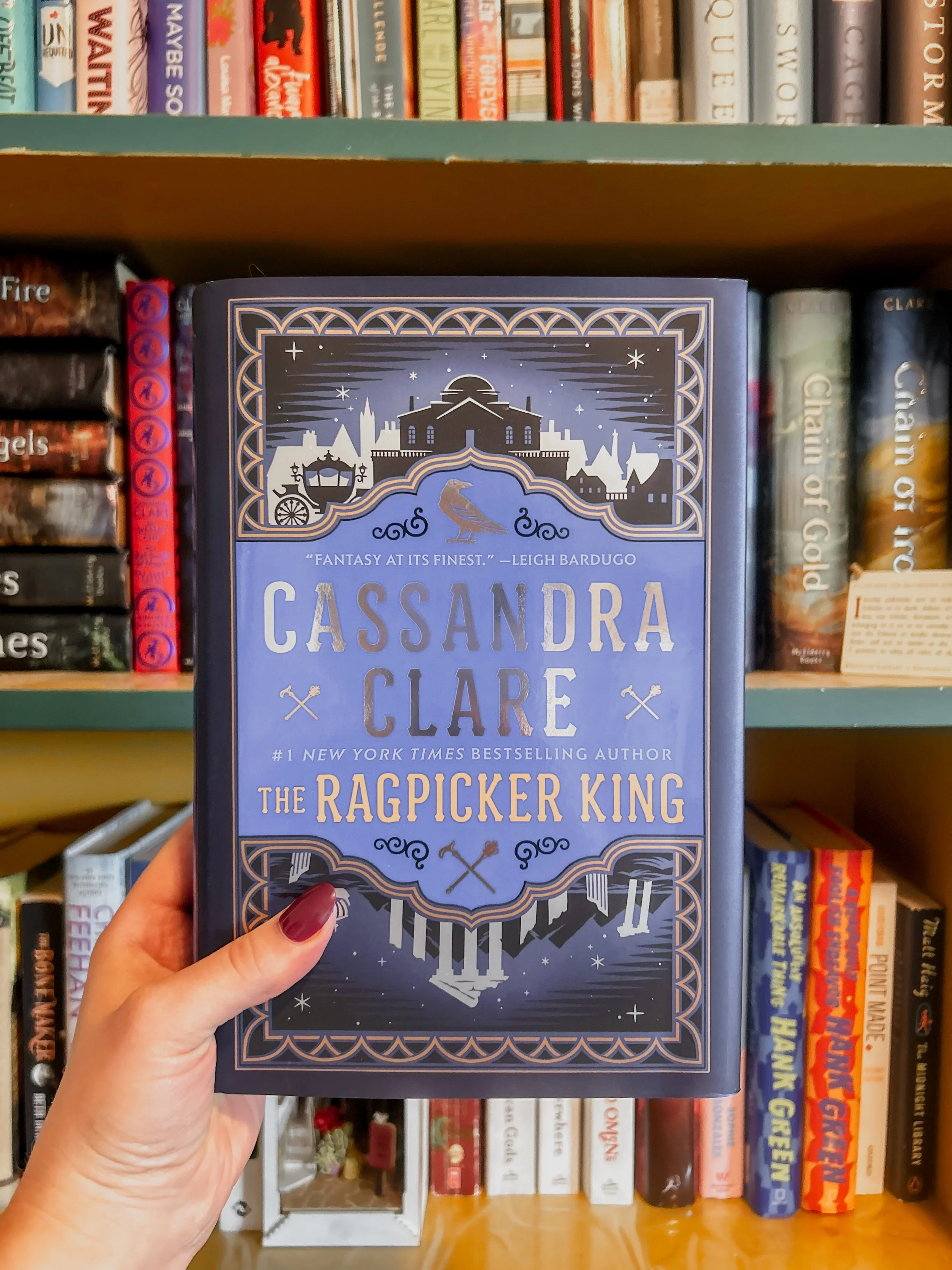 hand holds The Ragpicker King by Cassandra Clare in front of a colorful bookshelf