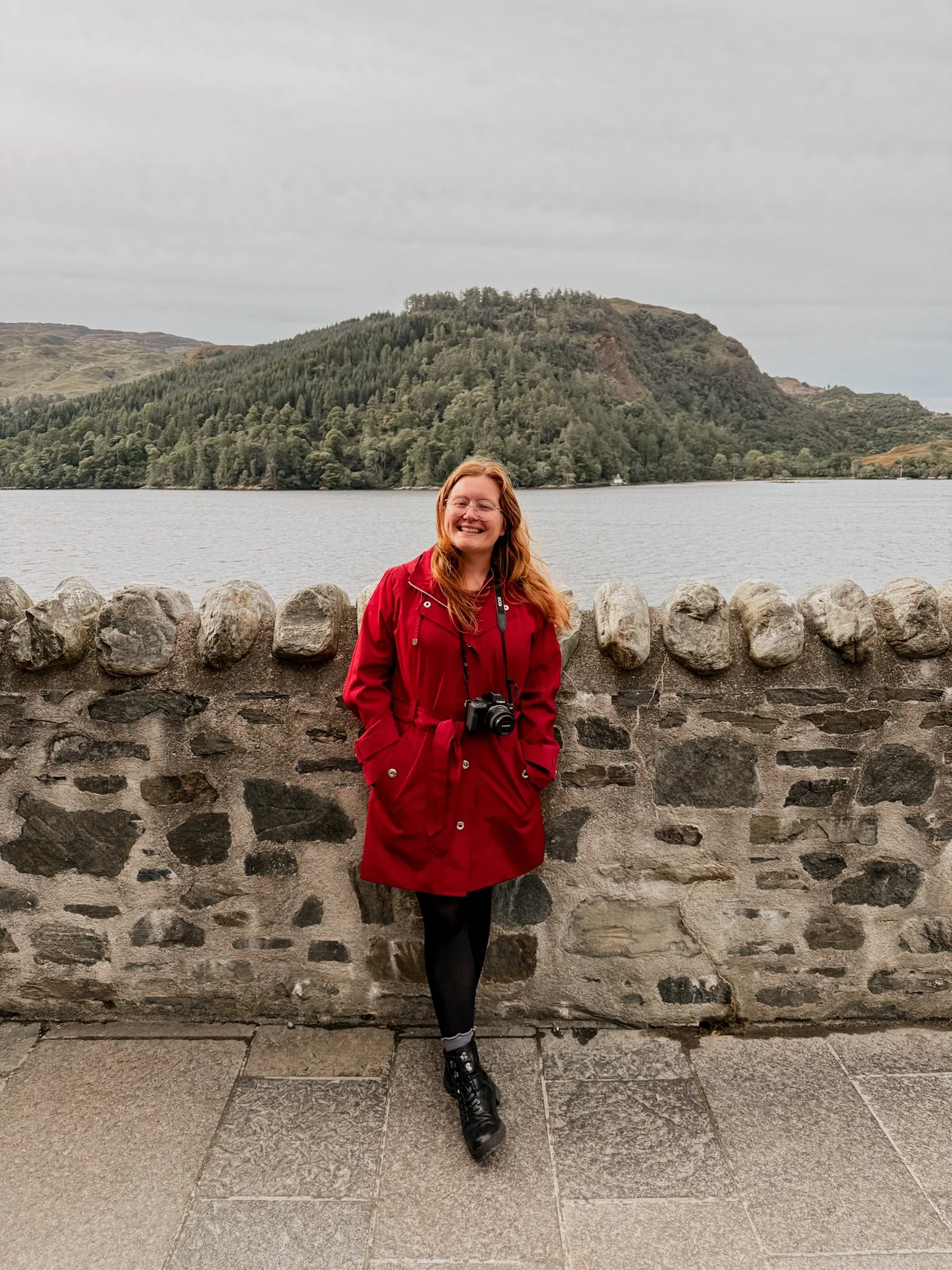 girl in red coat standing in front of stone wall and lake