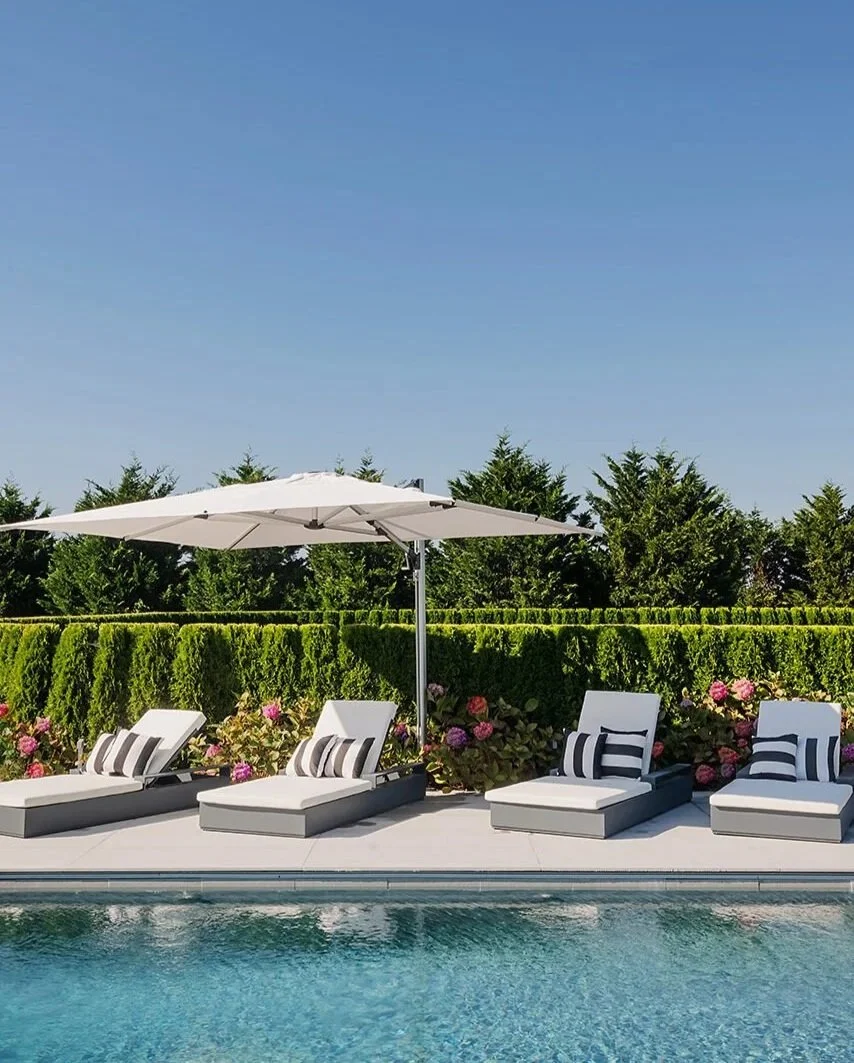Poolside scene with four lounge chairs, striped pillows, and a large white umbrella under a clear blue sky, surrounded by green hedges and flowering bushes.