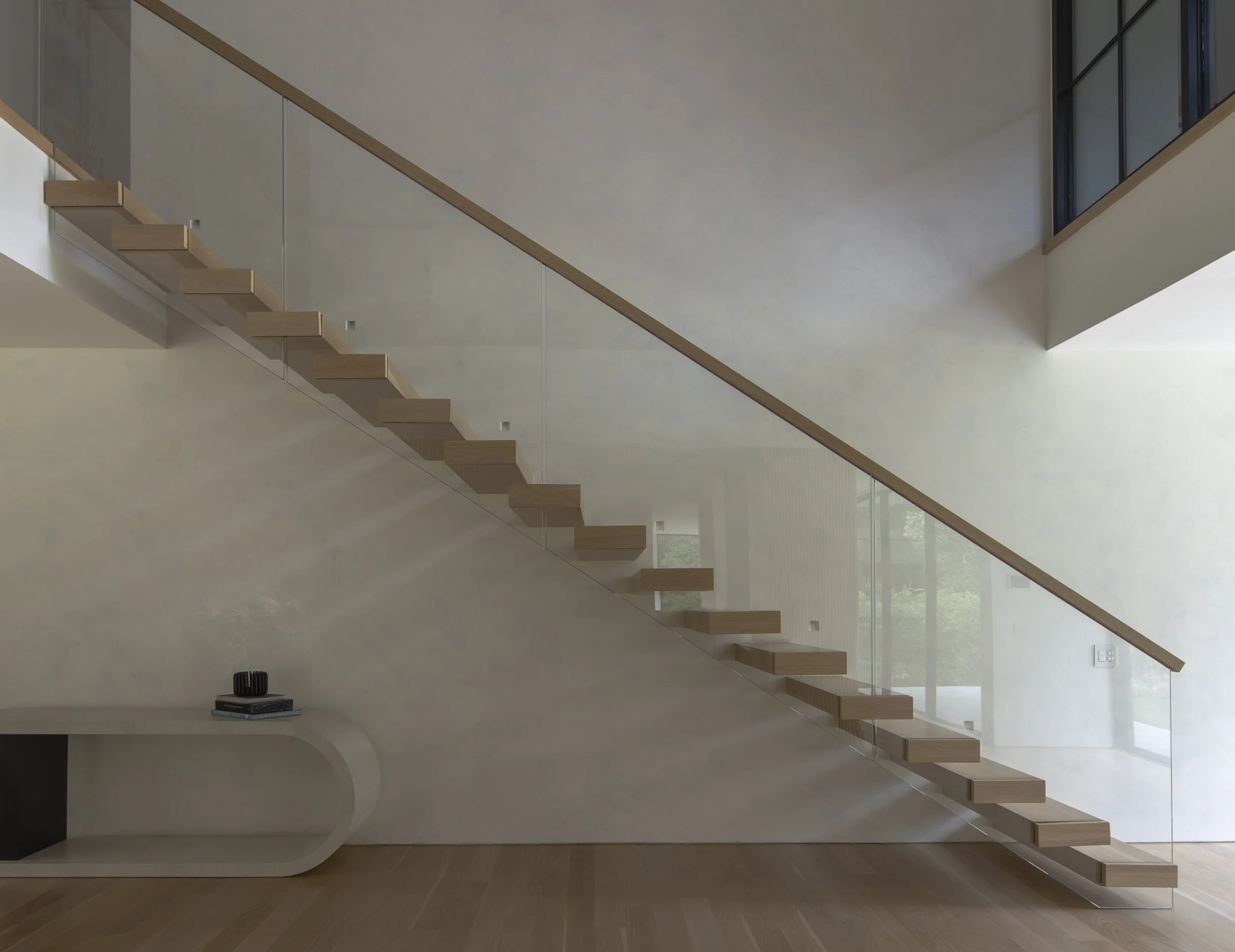 Interior view of a modern staircase with wooden steps and a glass railing in a spacious, minimalistic room with a large window and light-colored walls.