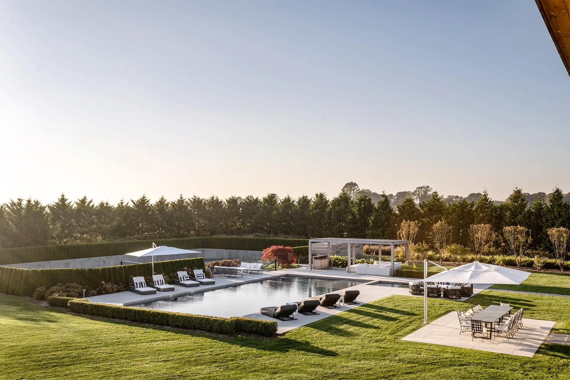 Luxury backyard with a rectangular swimming pool surrounded by gray tile deck, sun loungers, umbrellas, outdoor dining table, and lush green lawn, with trees in the background and a clear sky.