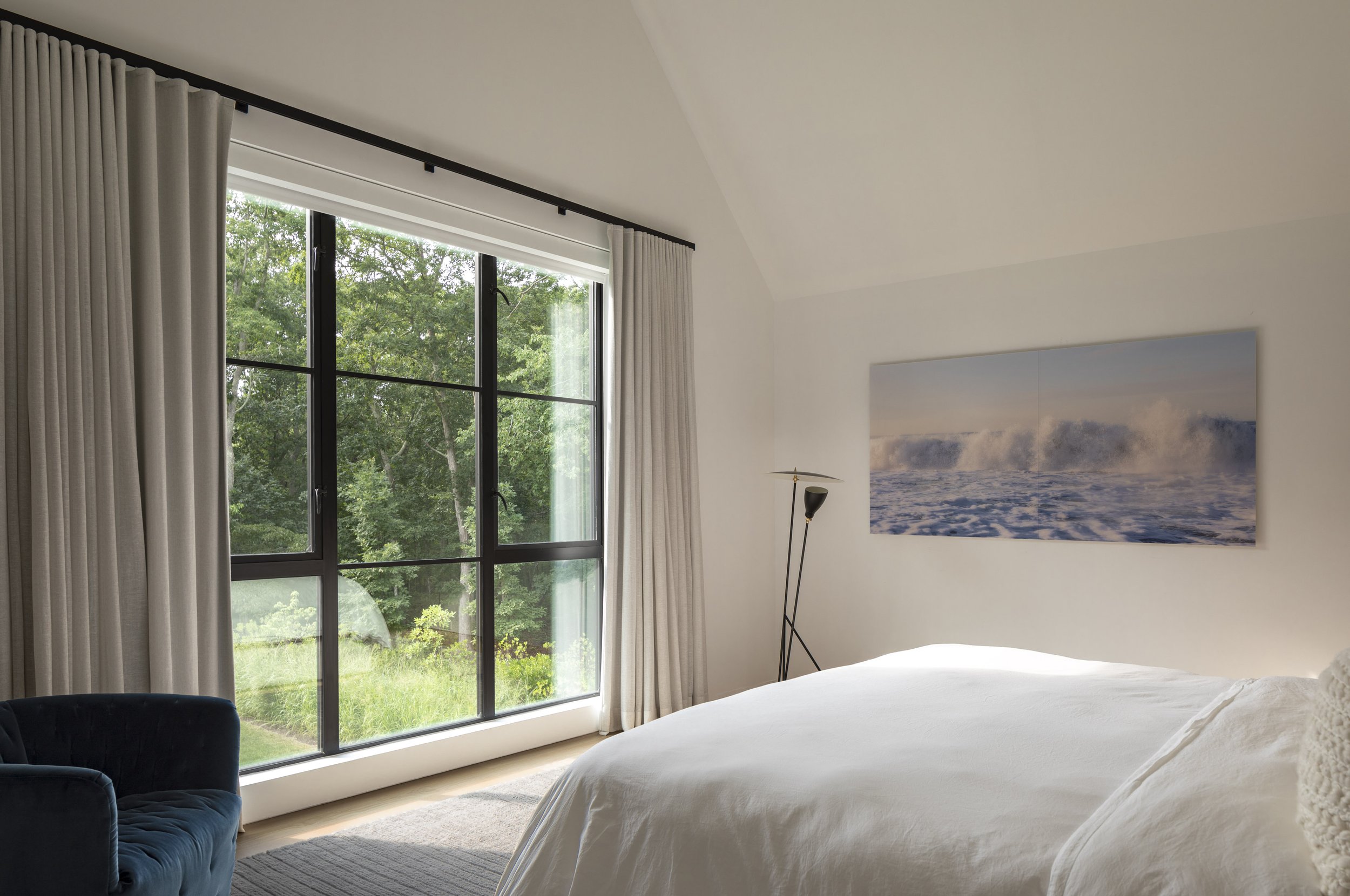 A bedroom with large window overlooking a green outdoor space, white walls, a bed with white bedding, a black floor lamp, a blue armchair, and a ocean wave artwork on the wall.