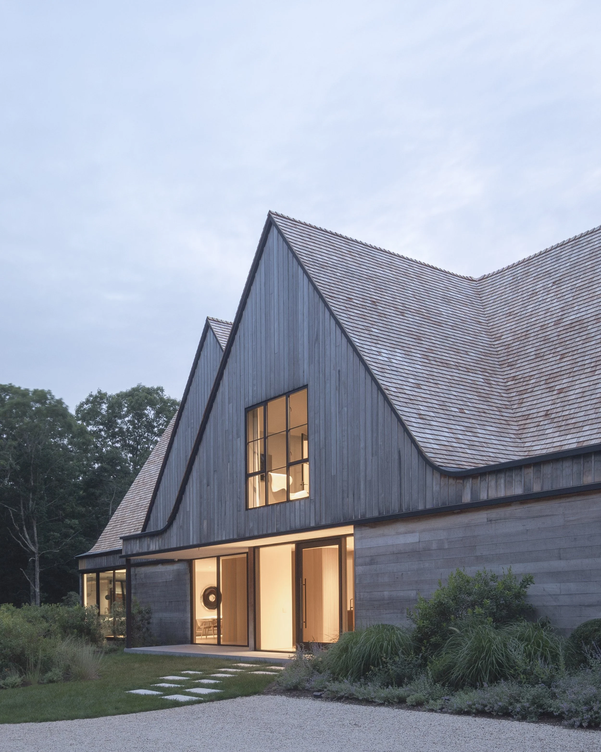 Modern house with a steep gabled roof, large windows, and a wood exterior. The porch area is lit from inside, and the house is surrounded by garden and trees under an overcast sky.