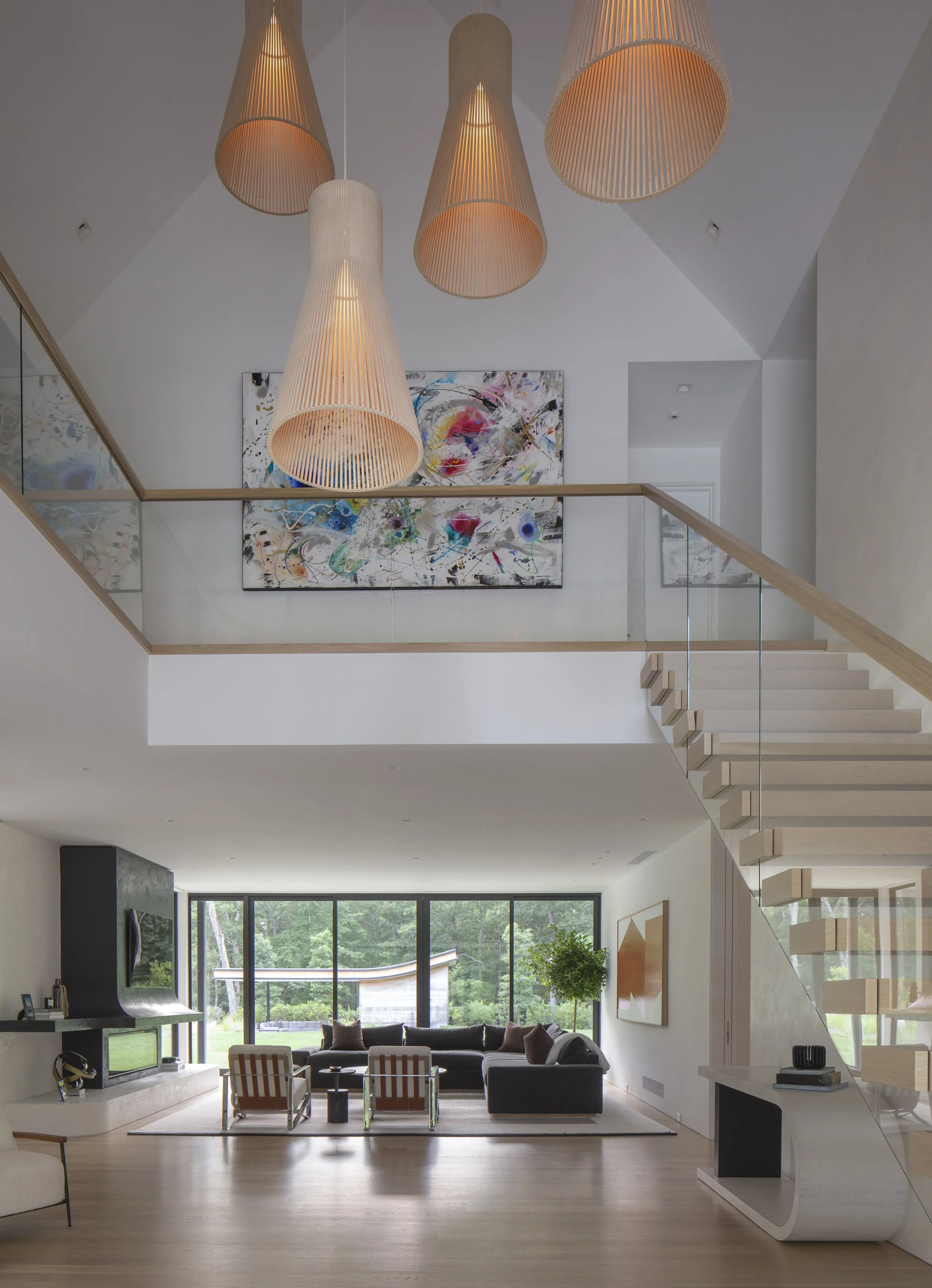 Modern living room with large windows, abstract artwork, and a staircase with wooden steps and glass railing.