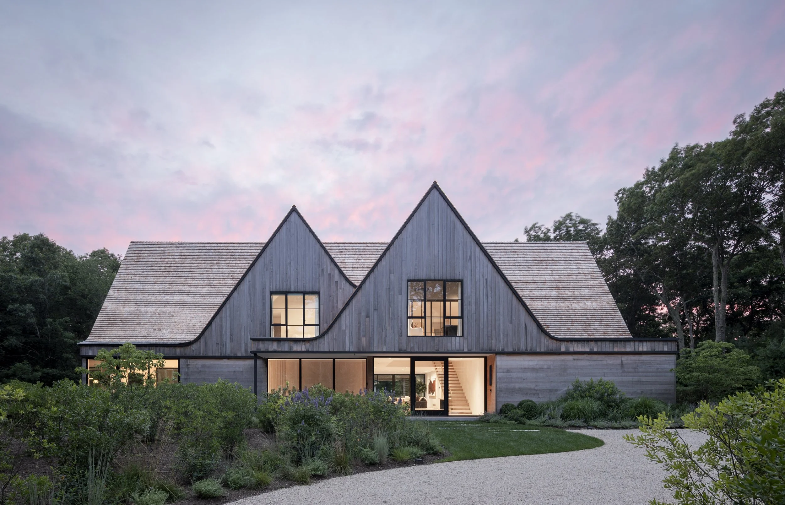 Modern house with wooden exterior, large windows, set in a landscaped garden with a gravel pathway, trees, and a pink and purple sky at dusk.