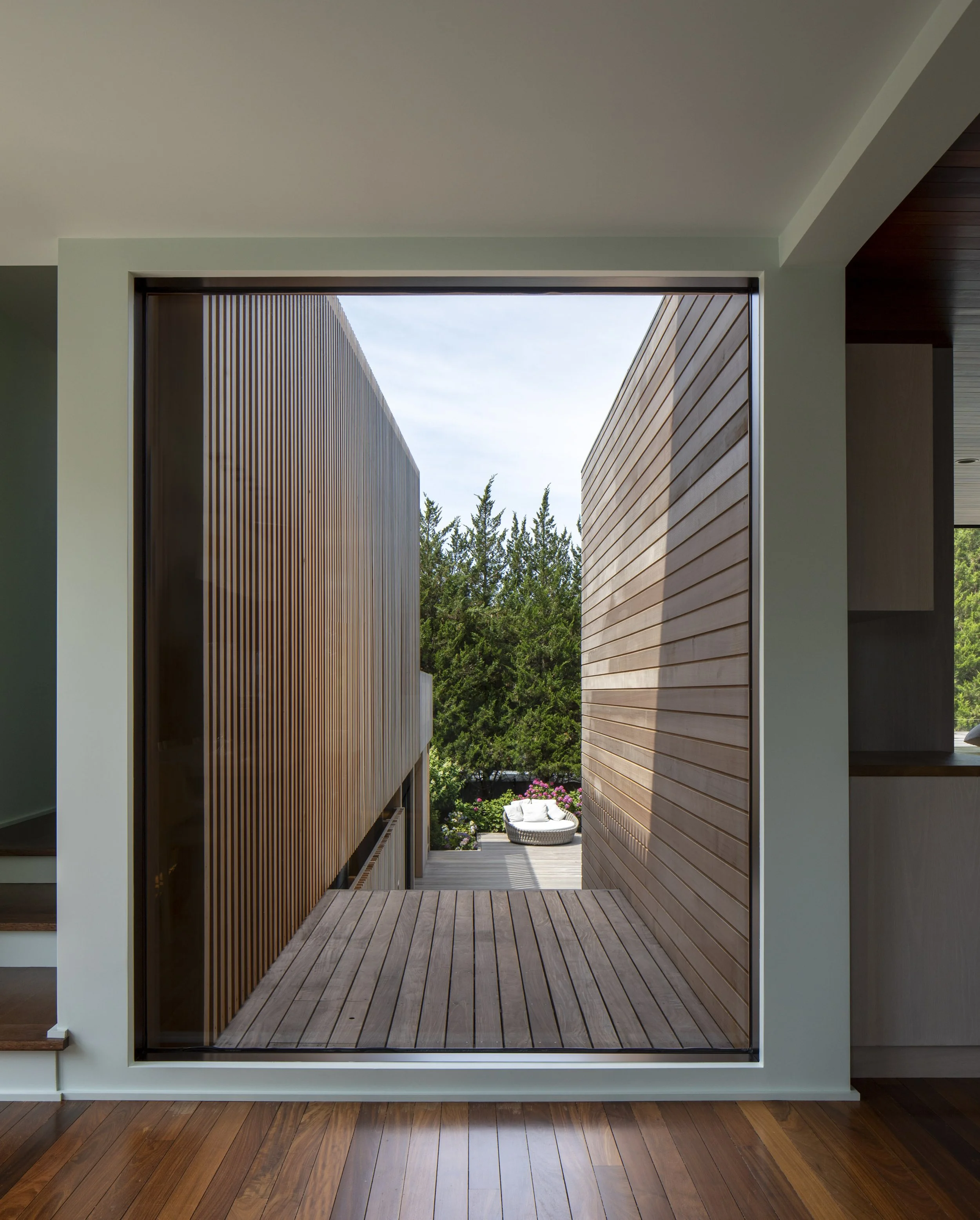 Interior view of a modern house with a large square opening framing a wooden deck and outdoor seating surrounded by trees and greenery.