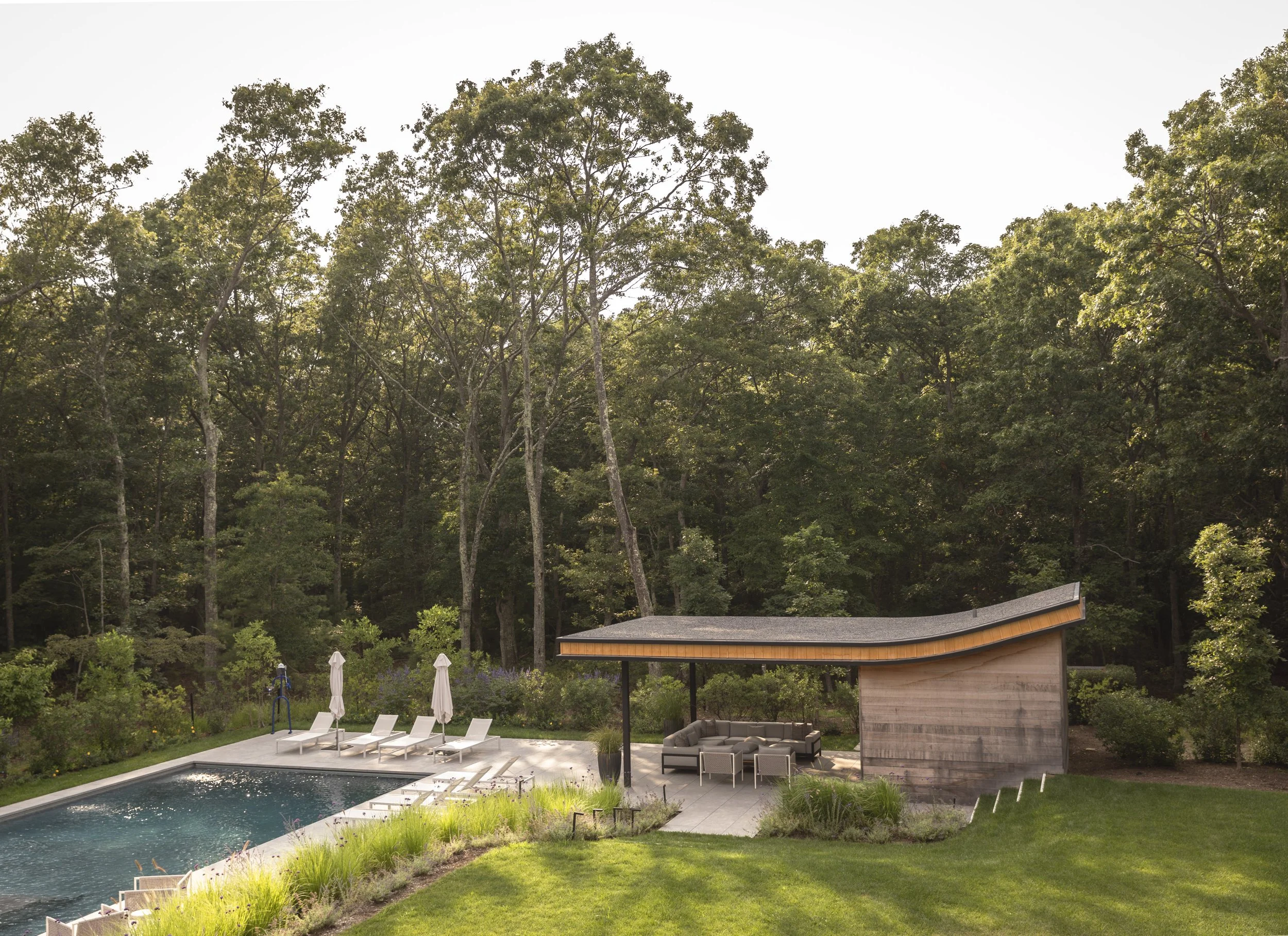 Backyard with swimming pool, poolside lounge chairs and umbrellas, outdoor seating area with sofa and chairs under a curved wooden roof, surrounded by green lawn and trees.