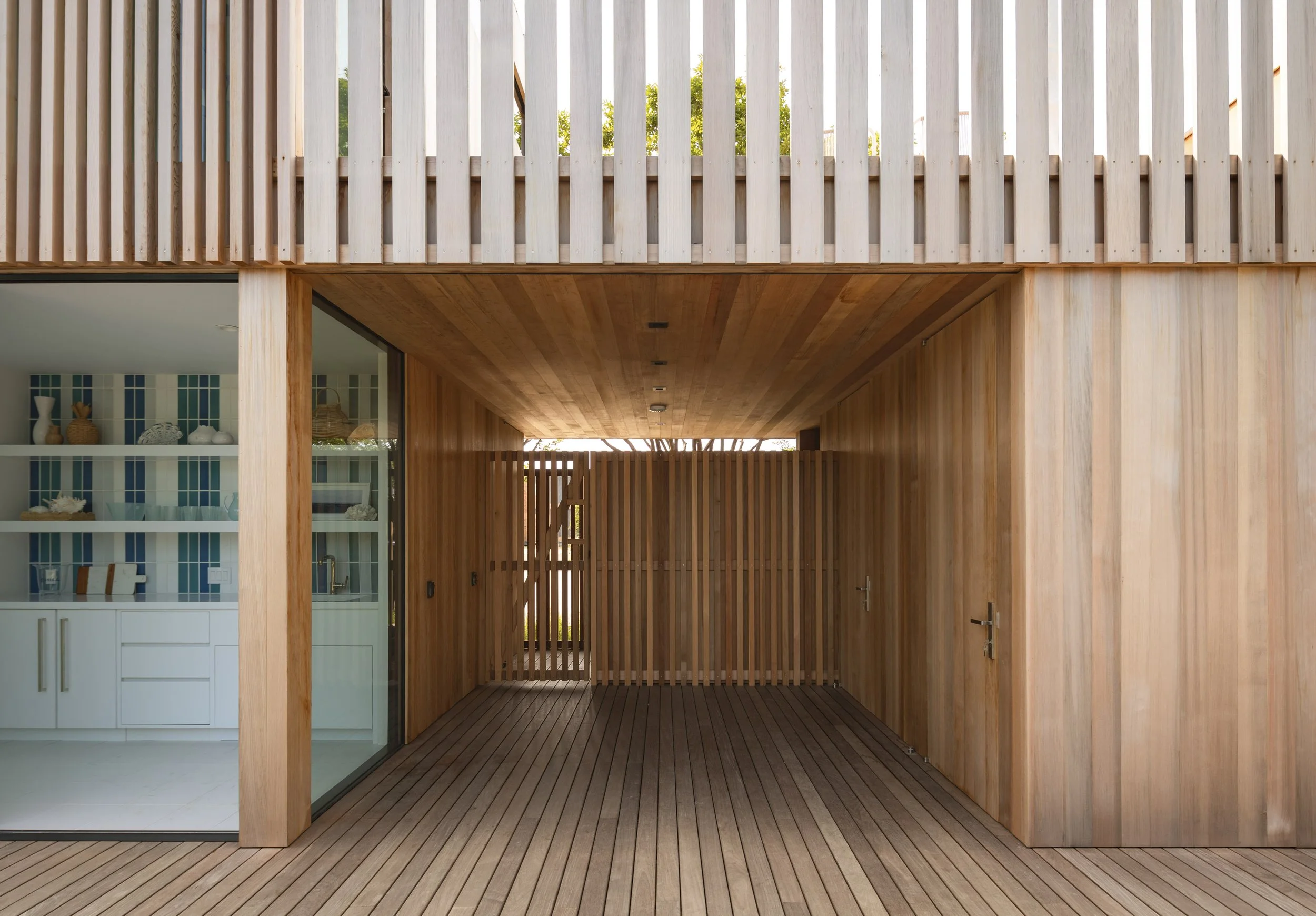 An outdoor wooden patio area with a wooden ceiling and walls, connected to a house with a glass sliding door revealing a kitchen inside. The patio has wooden flooring and a wooden fence at the back.