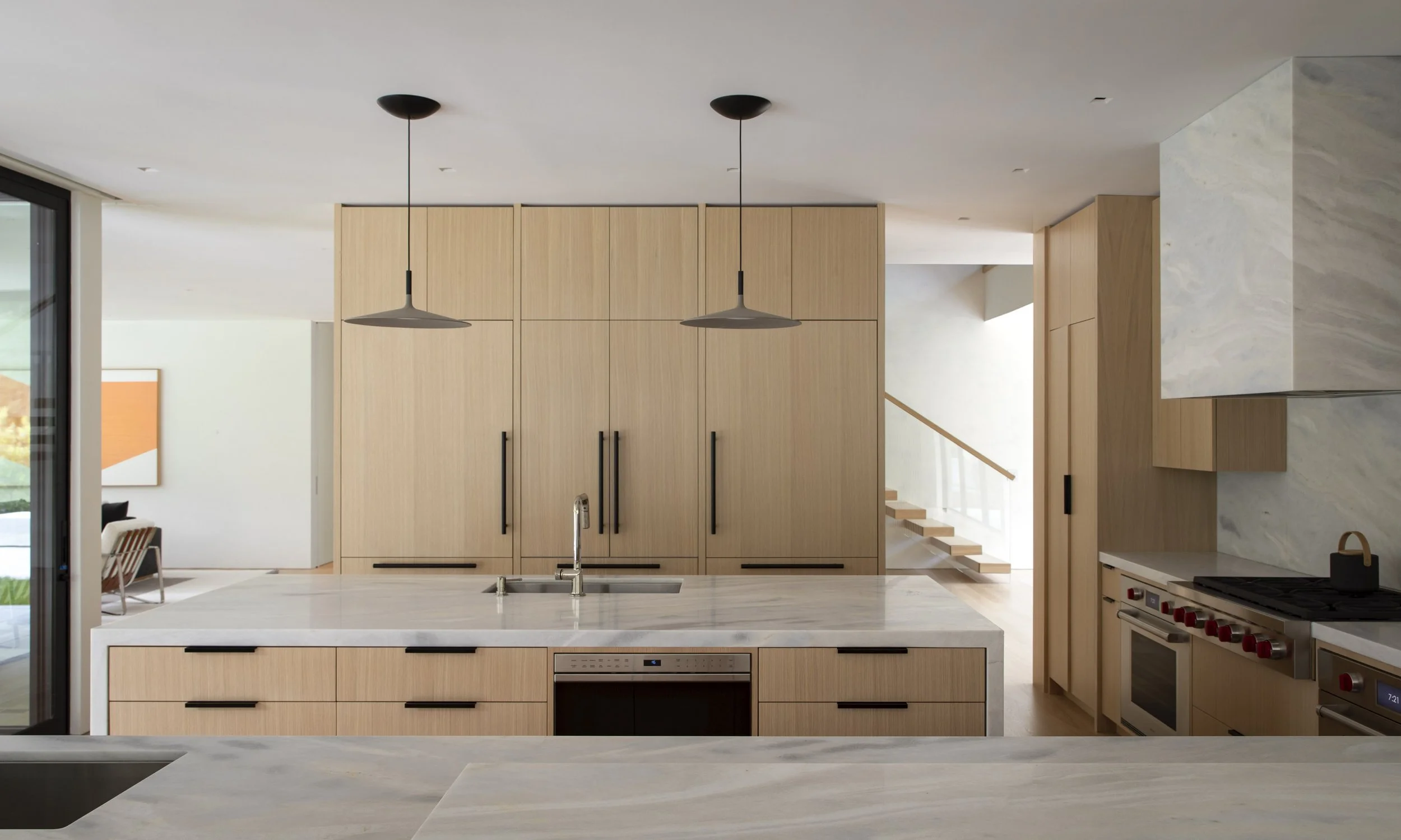 Modern kitchen interior with light wood cabinets, marble countertops, and black handles. Two pendant lights hanging from the ceiling, stainless steel sink, gas stove, and oven. Open space with a staircase in the background.