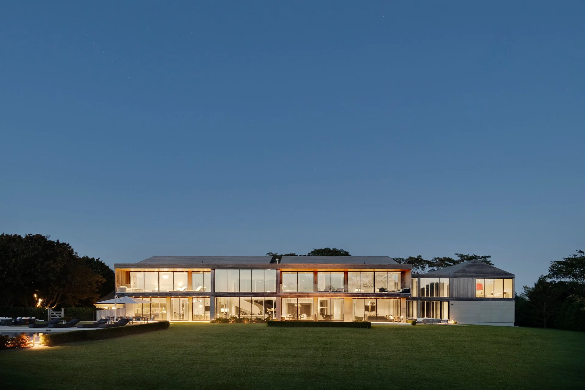 Modern two-story house with large glass windows illuminated at dusk, surrounded by a well-manicured lawn and trees in the background.