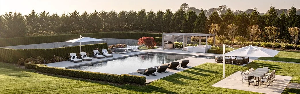 Luxury backyard with a rectangular swimming pool, lounge chairs by the pool, shaded seating area, outdoor dining table with chairs, and a well-maintained lawn and landscaping.