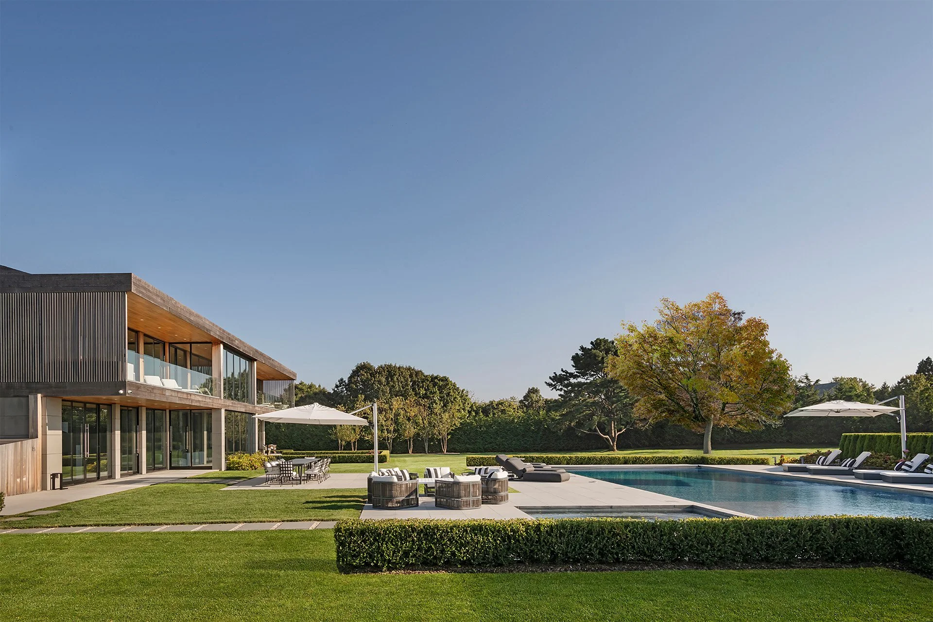 Modern house with large glass windows, outdoor pool, and patio furniture in a landscaped yard with trees and a clear blue sky.